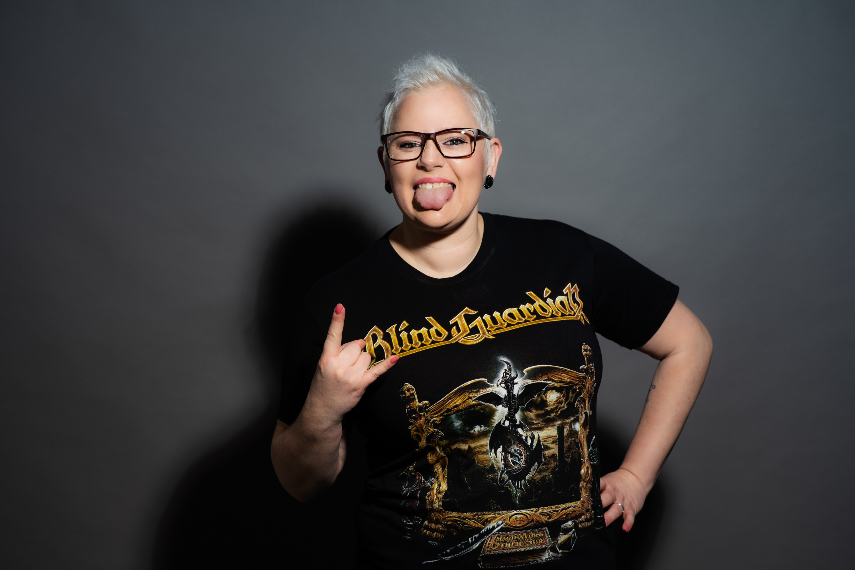 Studio photo of a woman with short platinum hair and glasses, playfully sticking out her tongue and flashing the rock hand sign, wearing a black “Blind Guardian” band T-shirt against a gray backdrop