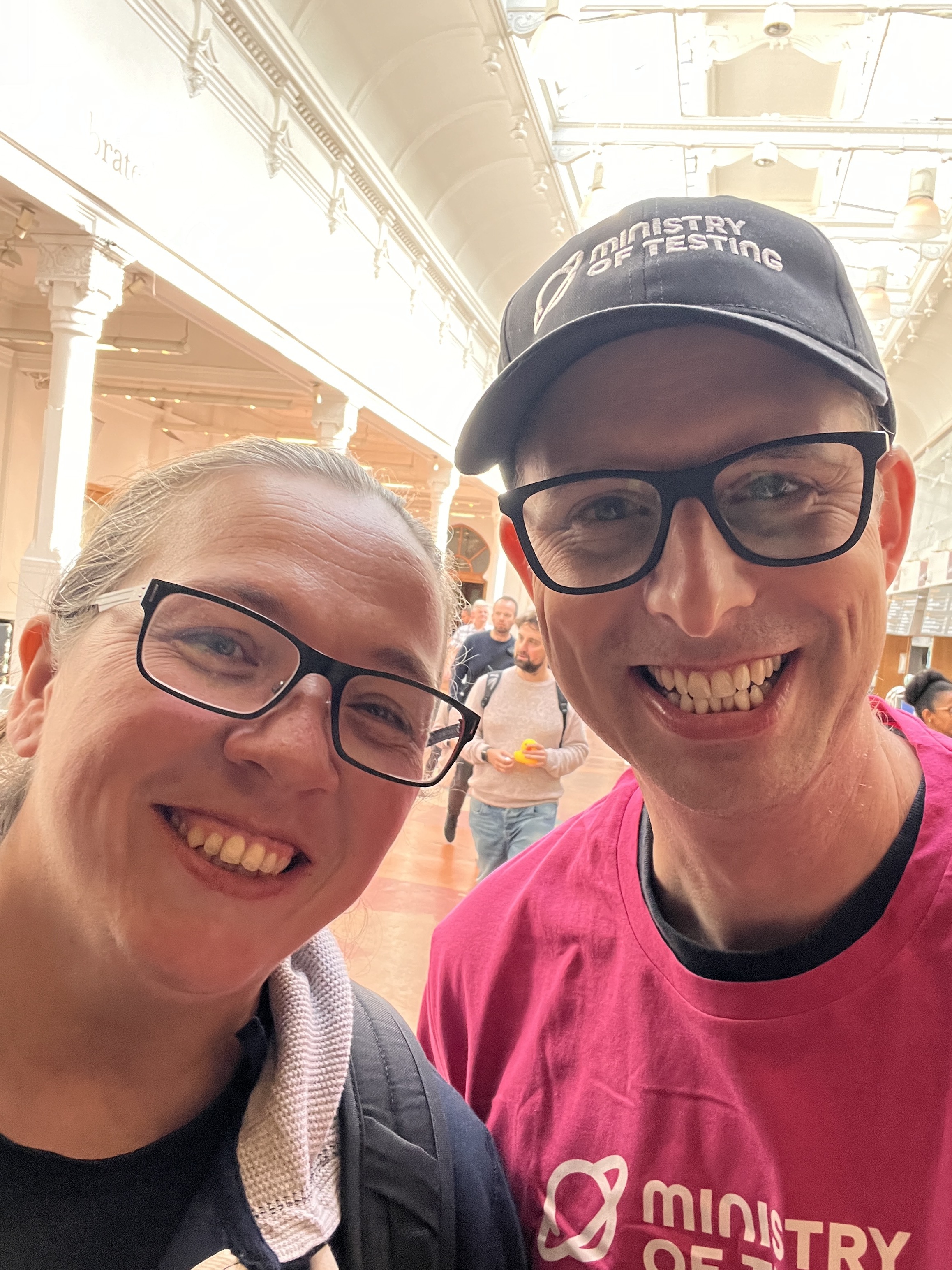 A smiling man and woman take a selfie indoors, with a crowd of people in the blurred background. The man wears a black baseball cap and a pink shirt, both with the "Ministry of Testing" logo.