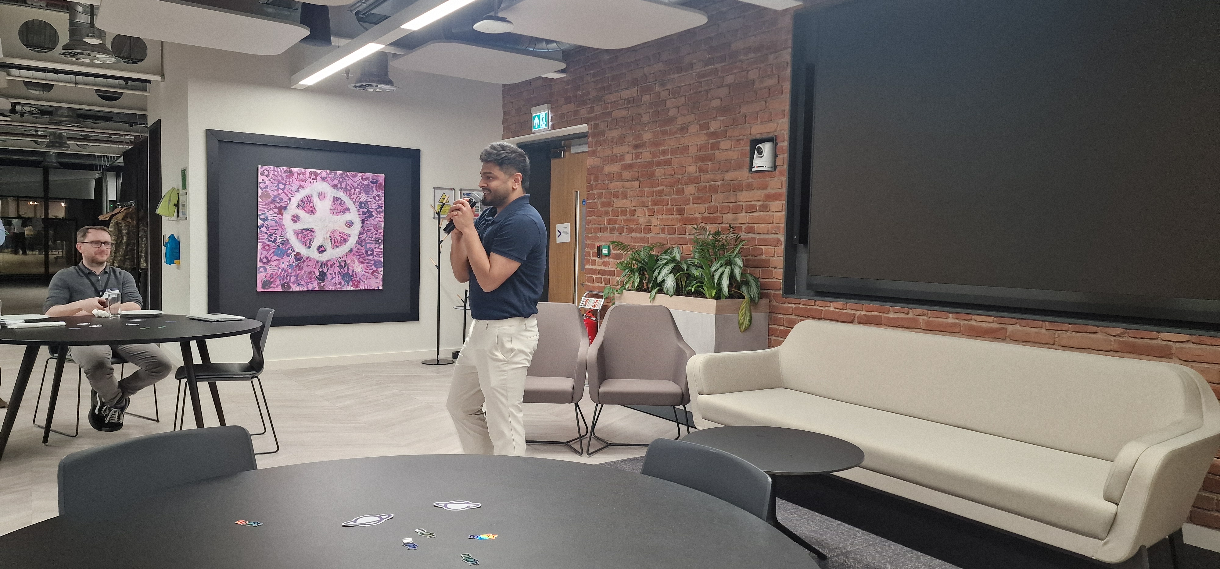 A person stands in the center of a modern office lounge, speaking into a microphone with both hands. They are wearing... image