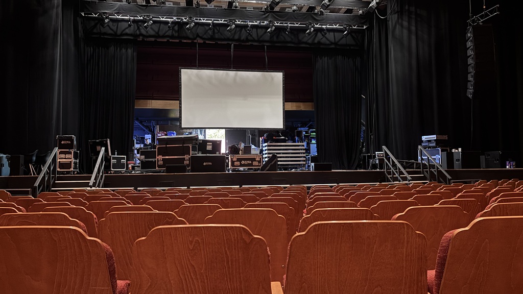 Rows of empty wooden seats facing the stage at Brighton Dome, with a projection screen, overhead lighting and equipme... image