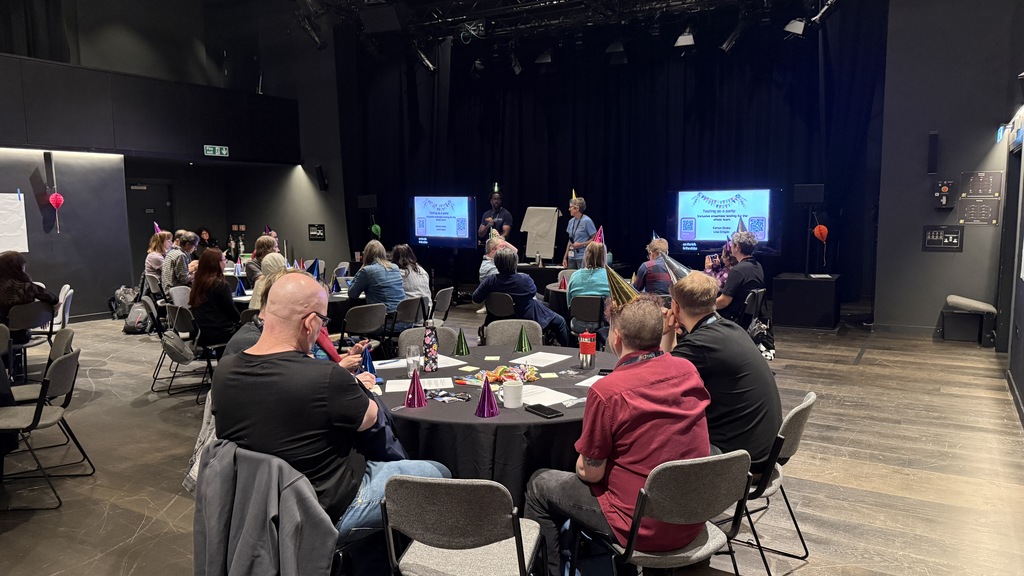 View from the back of the room for Lisa and Eamon’s workshop. People are sitting at round tables with the hosts at th... image