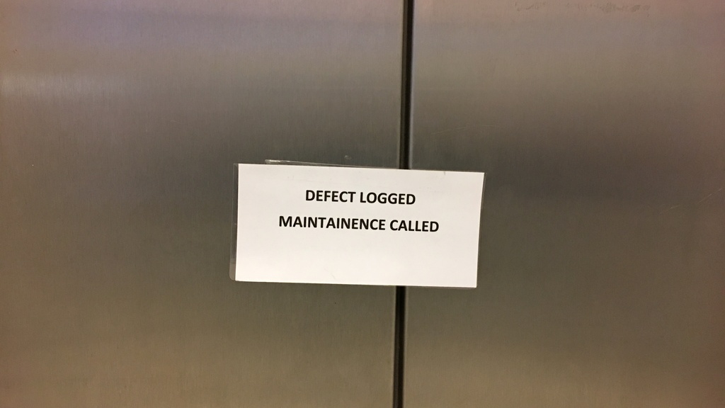 The image shows a printed sign taped to the closed doors of an elevator. The sign reads: "DEFECT LOGGED MAINTAINENCE CALLED." The word "MAINTAINENCE" is misspelled and should be "MAINTENANCE."