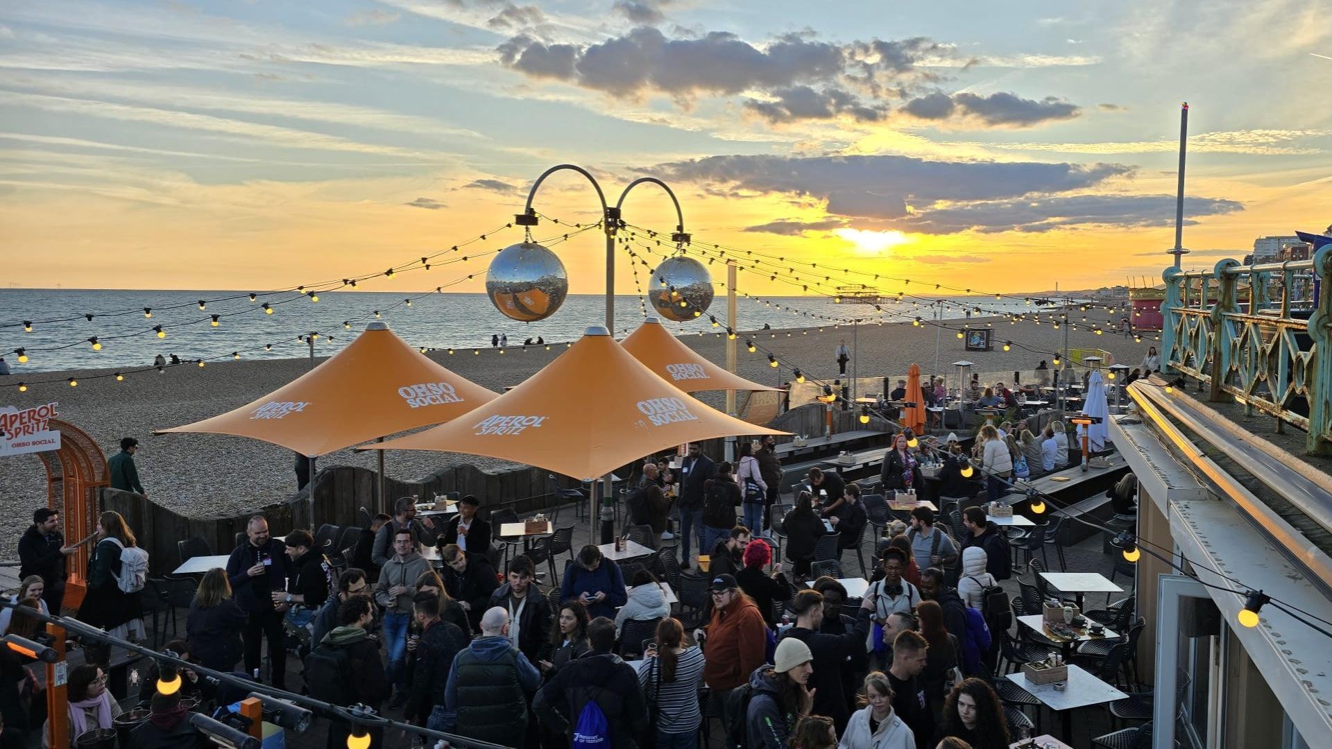 The sun sets over the sea. A large group of people are gathered in a social space on the beach. Many conversations are happening in different sized groups. There's a buzz in the air. There is a small bug character and a duck character immersed in the scene.