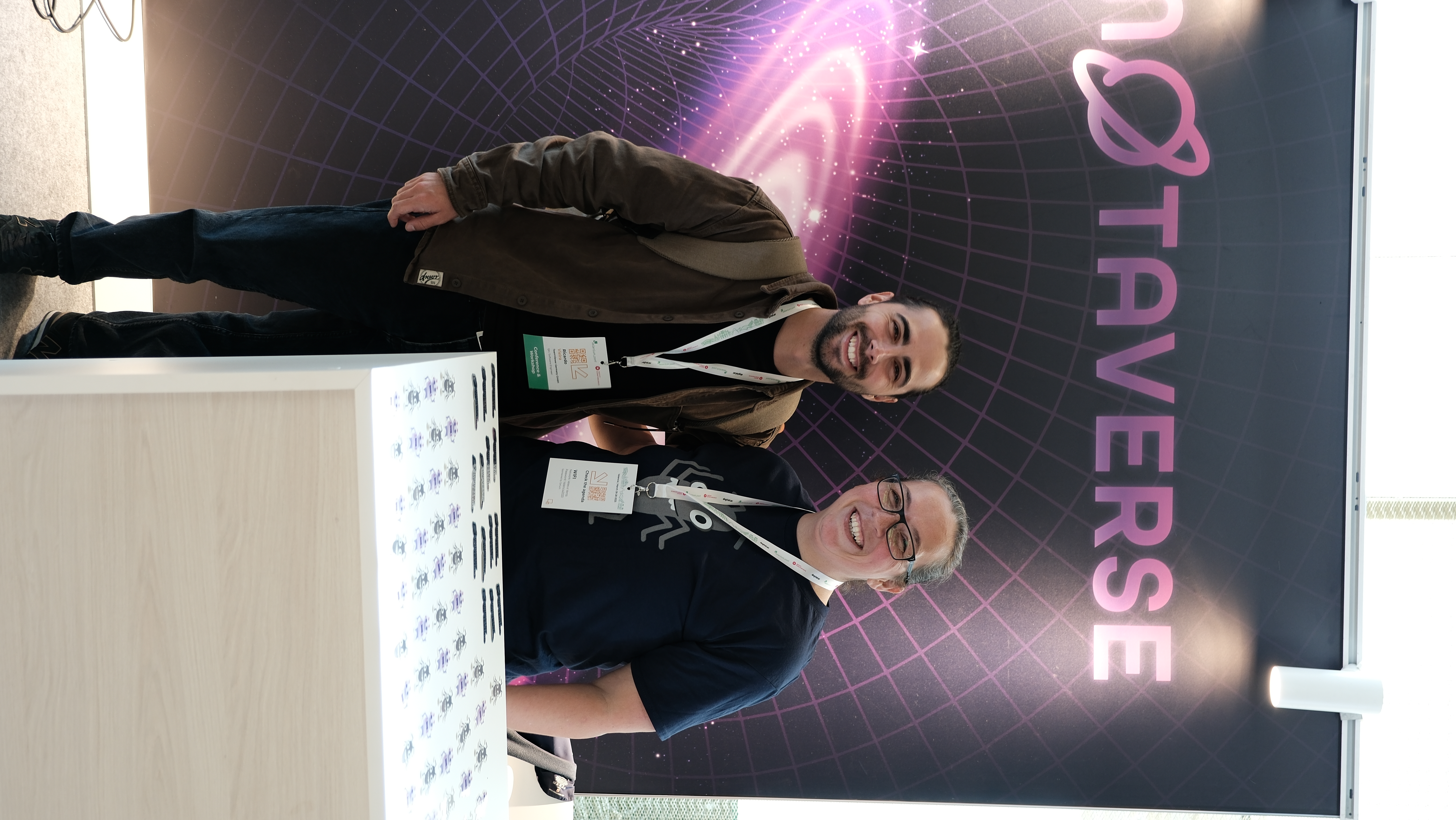 Ricardo Vieira stands smiling beside Rosie Sherry at the Ministry of Testing stand at Selenium Conf 2025, with the MoTaverse banner behind them and a table full of stickers in the foreground.