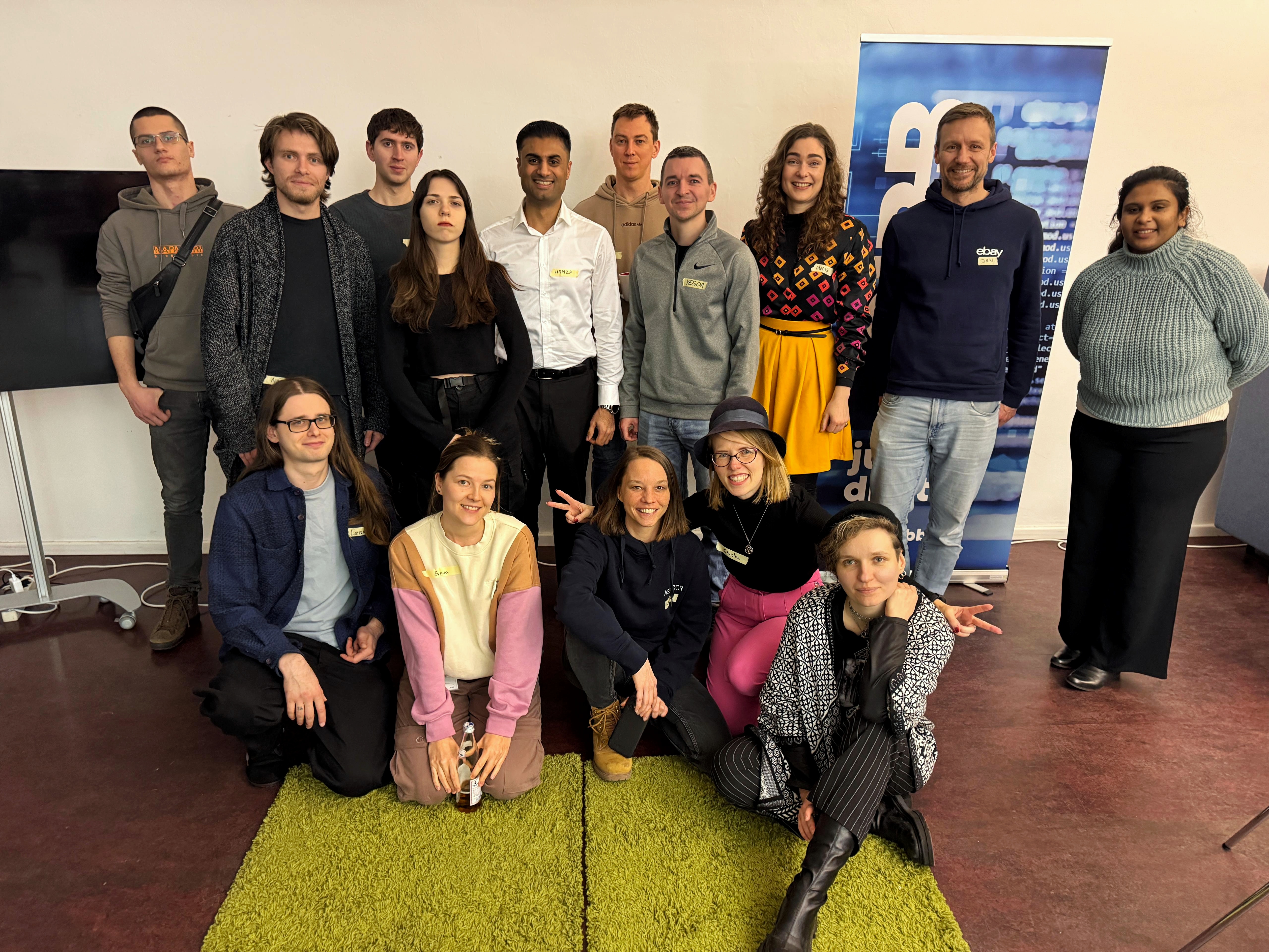 A group photo from a Ministry of Testing meetup in Berlin. The image shows 15 people arranged in two rows - 10 standing and 5 sitting/kneeling on a bright green carpet in the foreground. The group appears diverse and casual, with people wearing various informal attire like hoodies, sweaters, and casual business wear. In the background, there's a white wall with what appears to be a blue banner or poster. The atmosphere seems friendly and professional, typical of a tech community meetup."