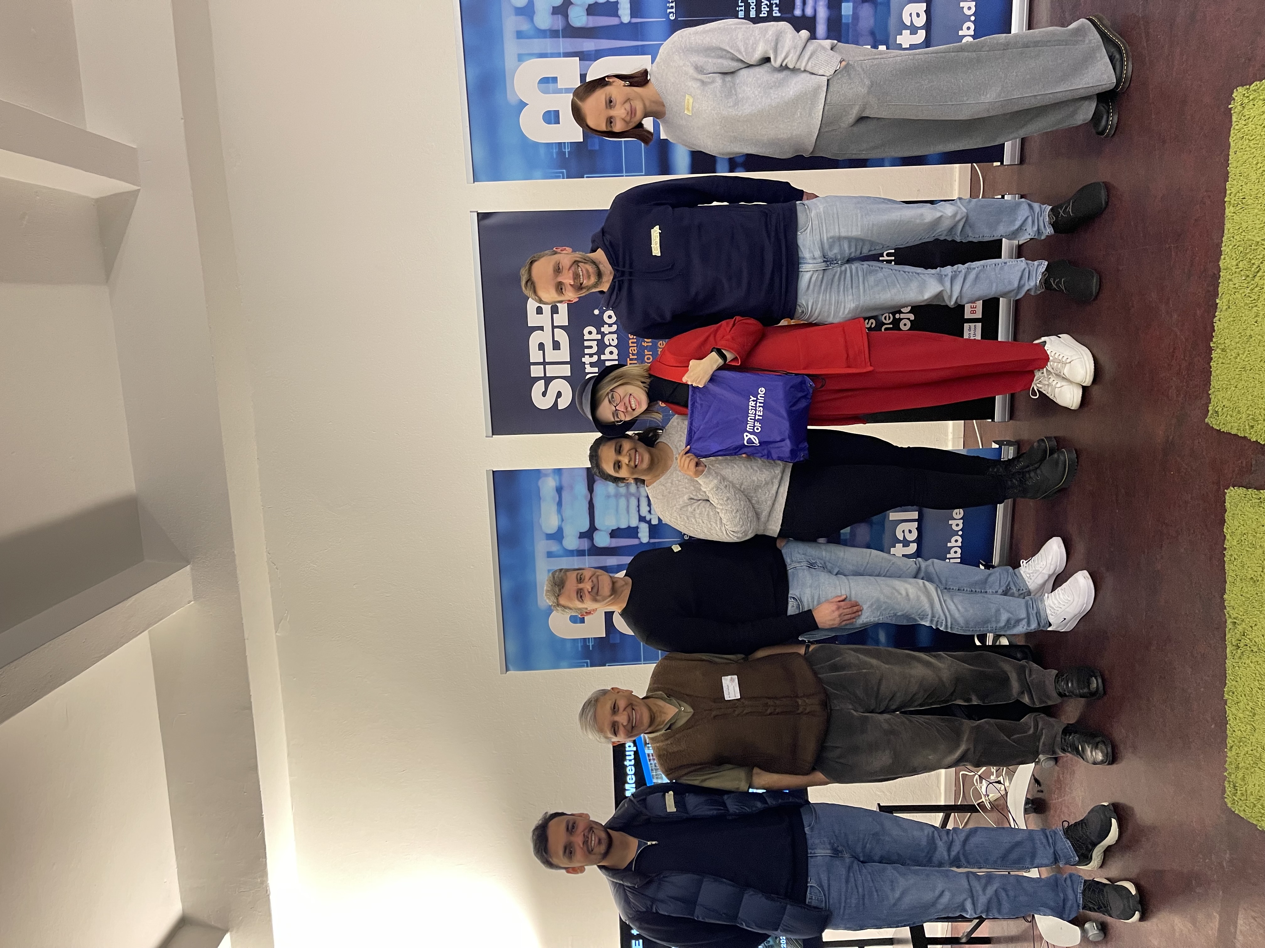 A group photo from the Ministry of Testing (MoT) Berlin meetup held at the SIBB Startup Incubator. Seven individuals are standing in front of banners with SIBB branding. One person holds a purple bag with the Ministry of Testing logo. Everyone is smiling in a casual and friendly atmosphere.