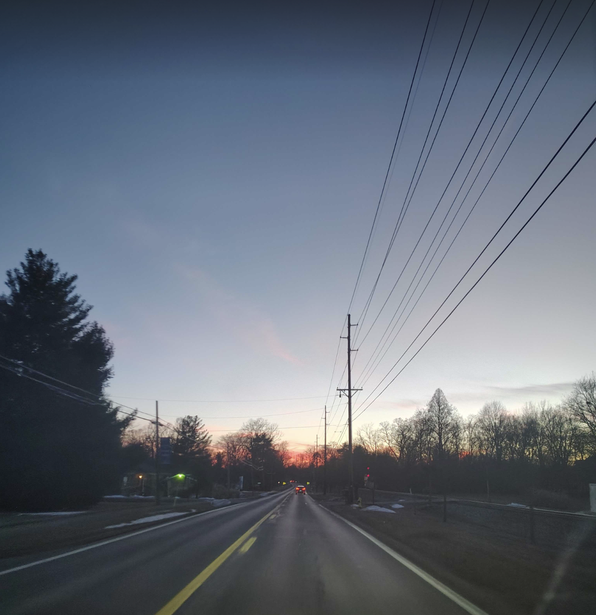 An image of a road set in twilight. The sun is setting. The trees are black against a pink sky. A car's taillights are glowing father ahead.