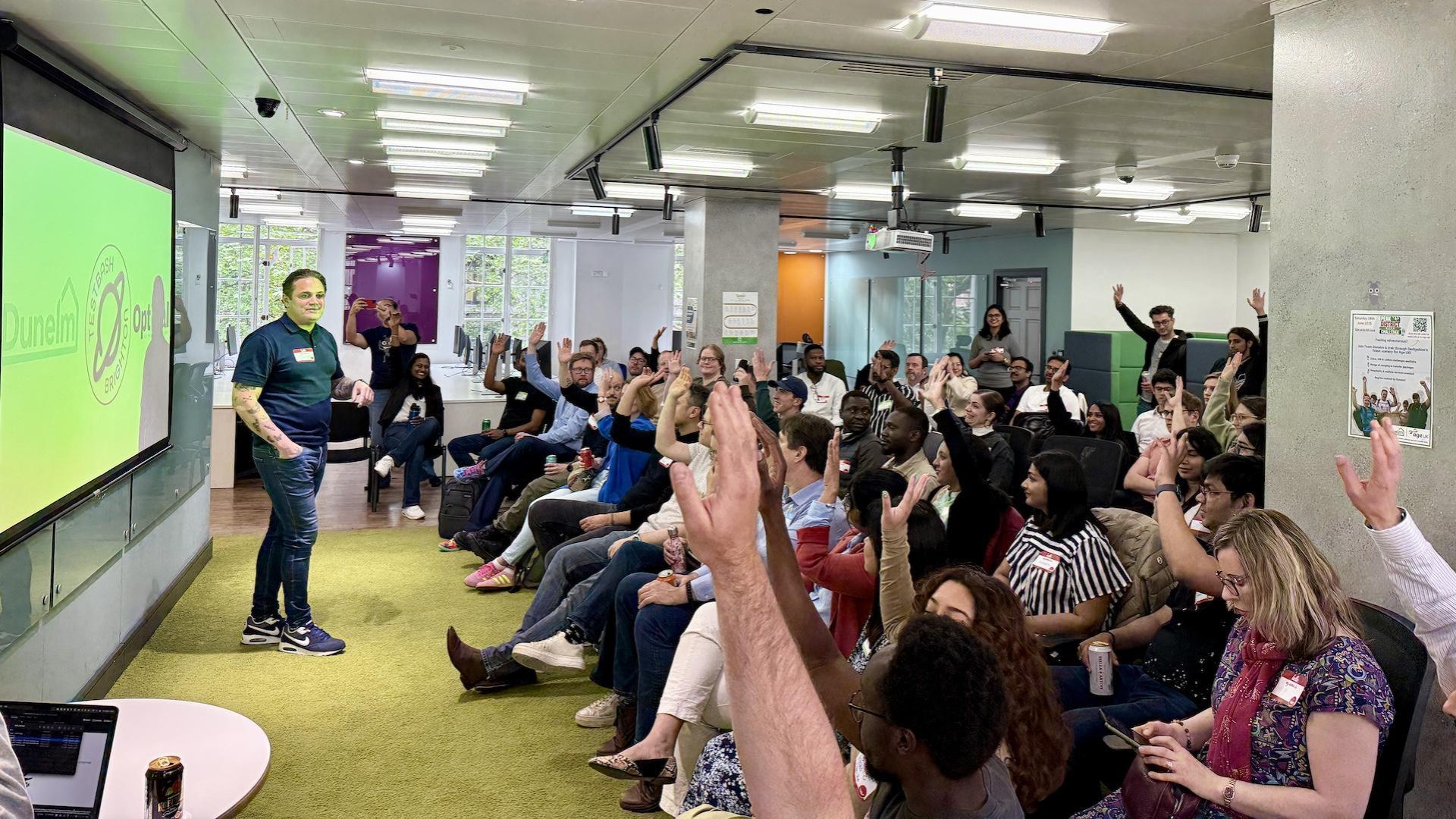 A man in a blue polo shirt and jeans stands to the left of a projection screen, addressing a large group of people seated in rows of chairs in a brightly lit room. Many of the seated individuals have their hands raised. The room has a modern design with fluorescent lighting fixtures on the ceiling, and glimpses of the outside greenery can be seen through windows along one wall. A few people are standing in the background near a doorway.
