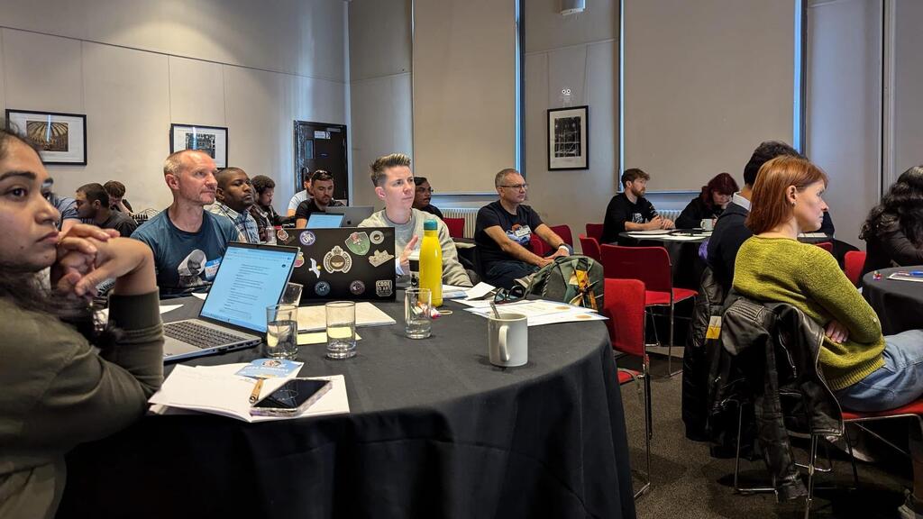  A diverse group of adults are seated at round tables in a conference room, engaged in what appears to be a training or workshop session. In the foreground, a woman on the left with dark hair is looking towards the front of the room, with her hands clasped. To her right, a man with short grey hair and a beard is also looking forward, with a laptop open in front of him. Another person with short, light-colored hair is seated further back at the same table, looking towards the front. On the far right, another woman with reddish-brown hair, wearing a green knitted sweater, is looking to the left. Most individuals have laptops or notebooks open, suggesting a learning environment. The tables are covered with black cloths and have water glasses and mugs on them. In the background, other attendees are visible, some looking at their screens, others at the front. The room has light-colored walls with framed pictures, and there are red chairs around the tables. A bug, duck and seagull character have been added to the image. 