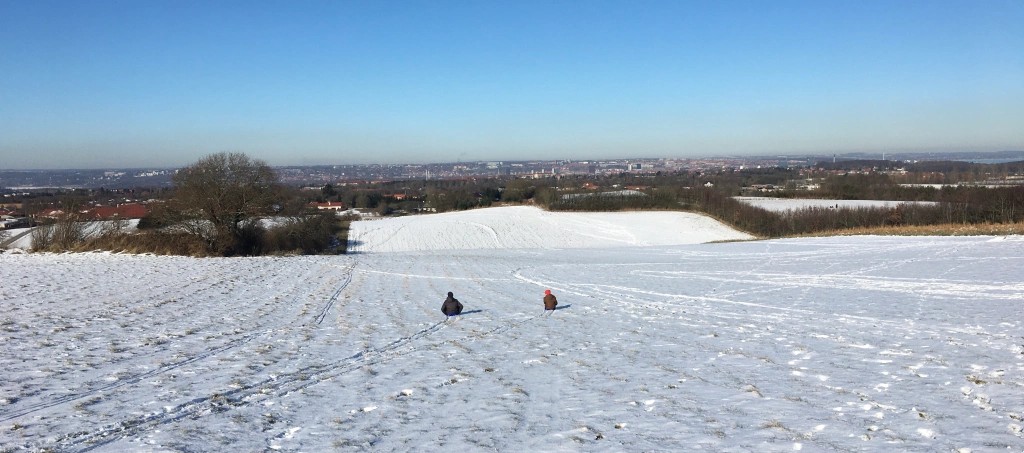 A snowy landscape slope with a view of a city in the distance. Two figures on sledges are going down hill. Photo taken some distance from them