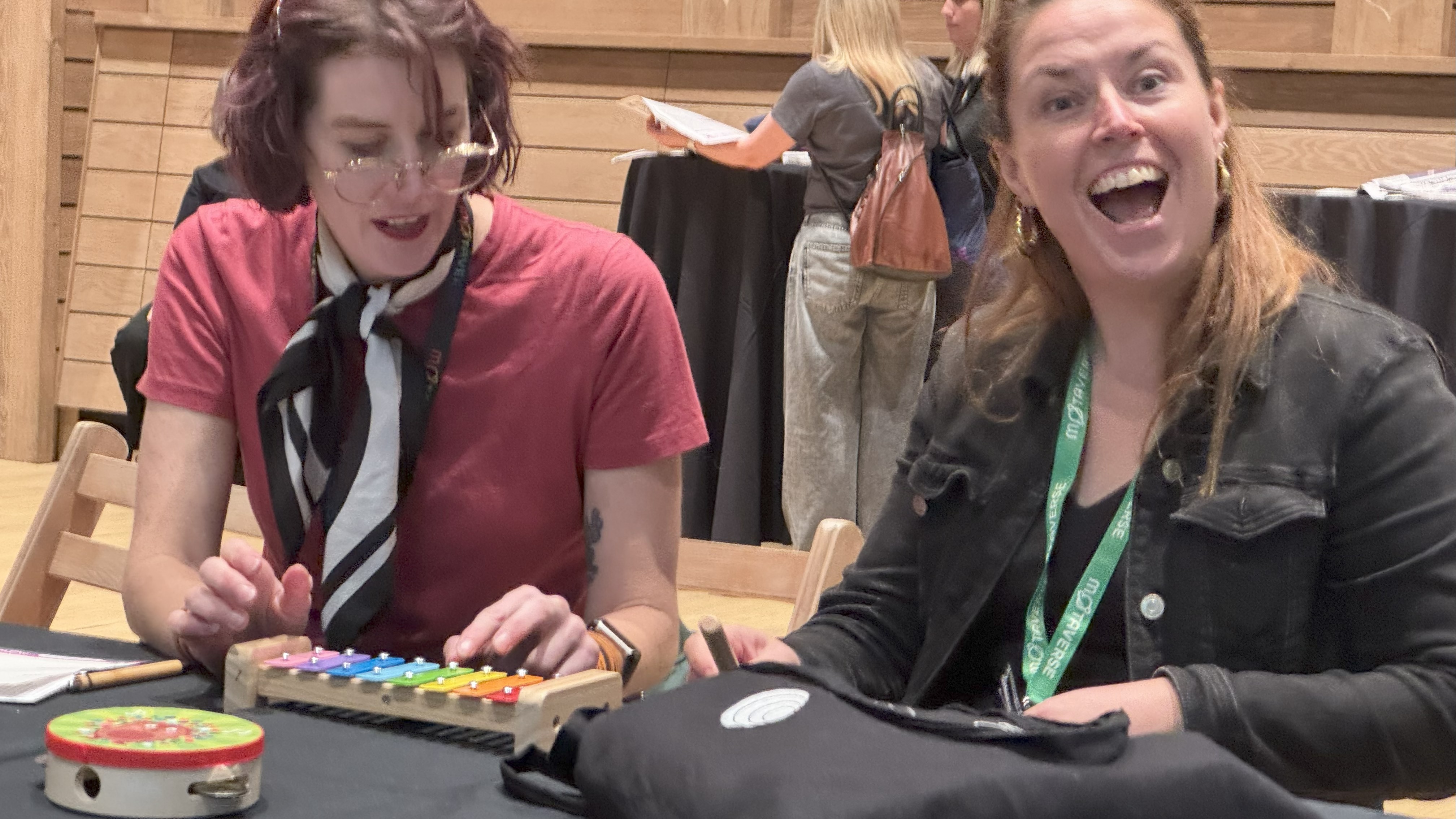 Gwen and Veerle are seated at a table, smiling and having fun. Gwen, on the left, is playing a colorful glockenspiel, while Veerle, on the right, grins widely at the camera. A tambourine and a black bag with a white logo lie on the table. The background shows other people standing and chatting in a wooden hall.