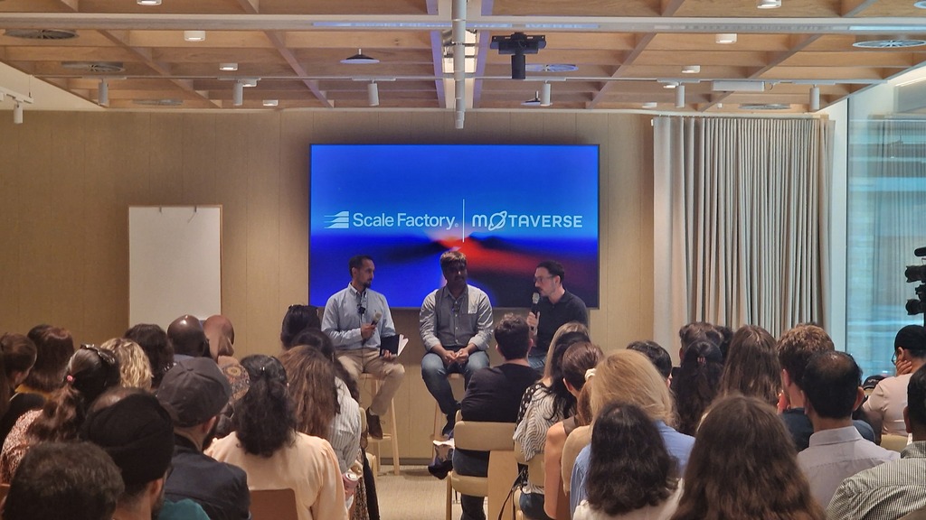 A panel discussion featuring three speakers seated on stools facing a large audience. Behind the panel, a large scree... image