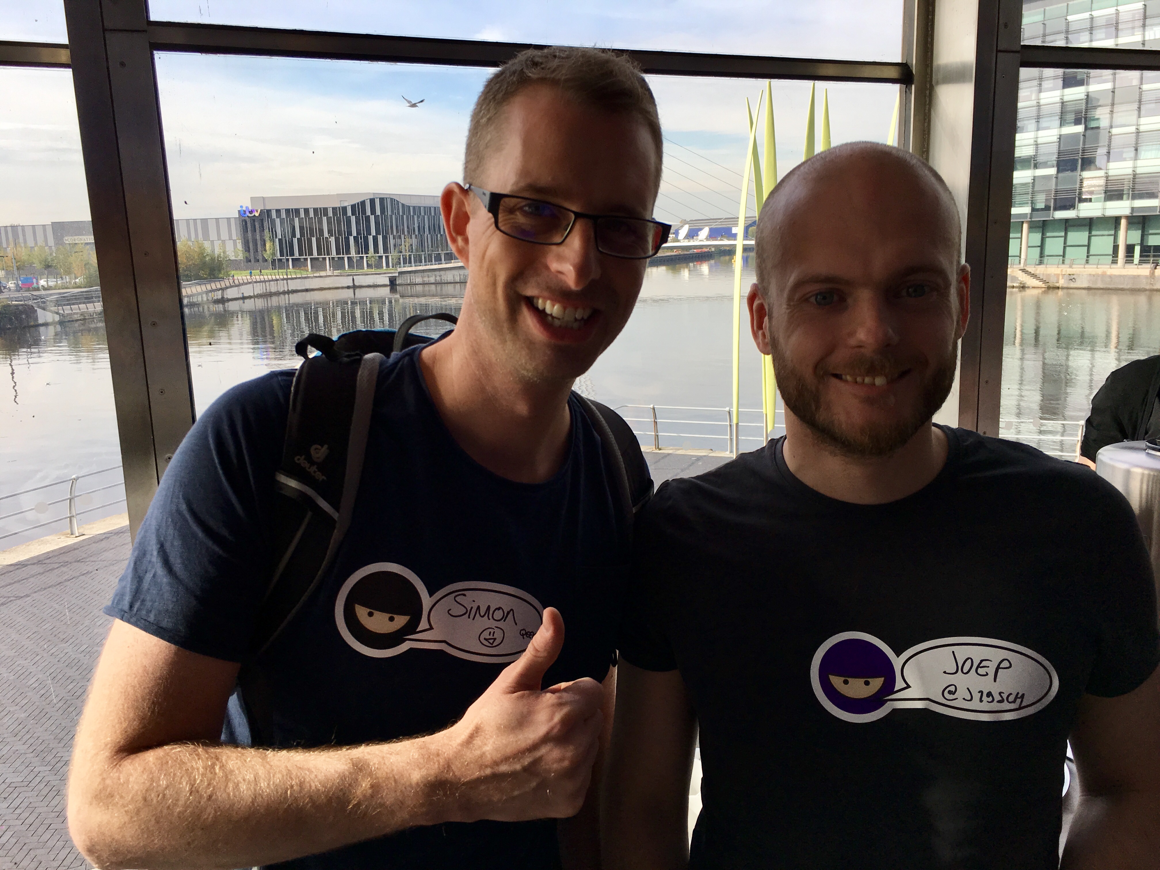A selfie of two white men in an event space. There is a water in the background. 