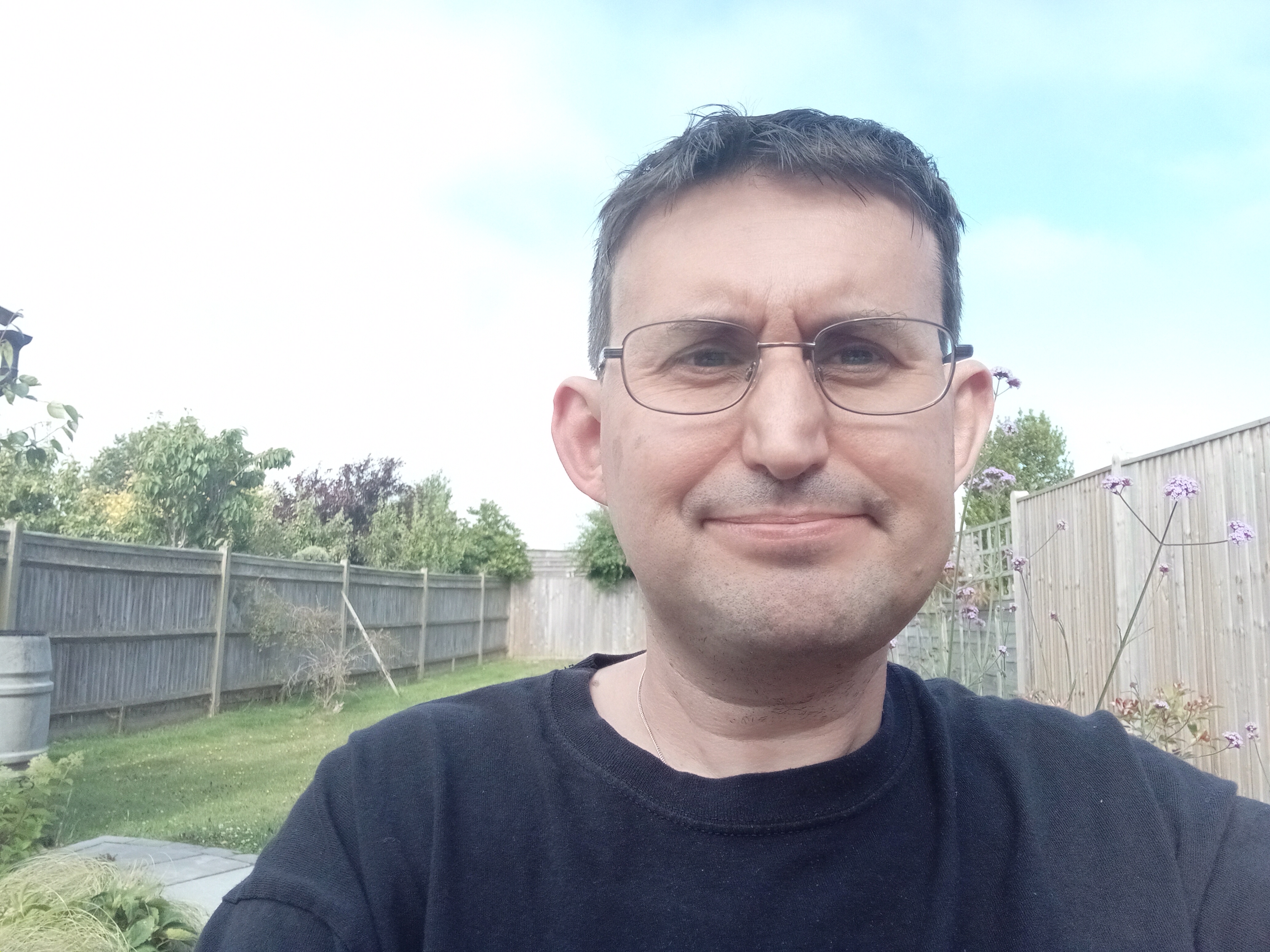White male with short black hair, wearing glasses, a black t-shirt and sitting in a garden.  The garden is surrounded by fencing, with green glass.  To the right in the picture is a plant with light purple flowers.