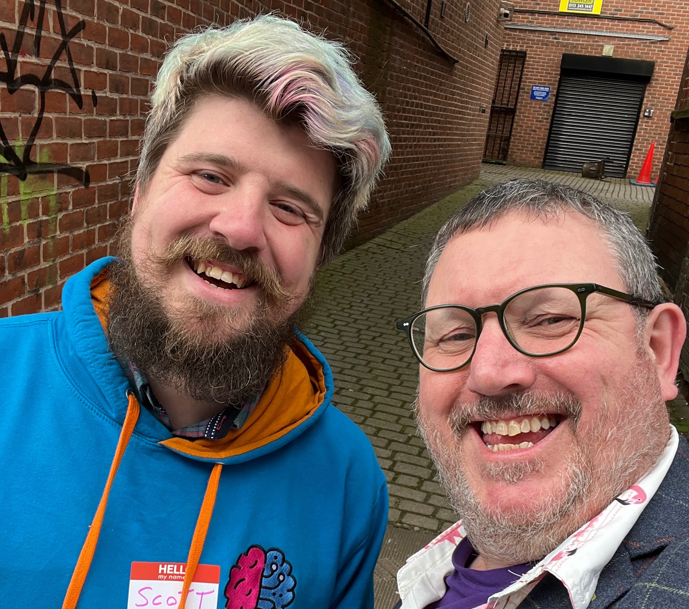 Me and Scott Kenyon outside Test Atelier in Leeds. Scott has a big beard and bright blond hair with a touch of pink. He’s wearing a bright blue hoodie. I have a stubble beard, glasses and I’m wearing a Hawaiian shirt and checked blazer. 