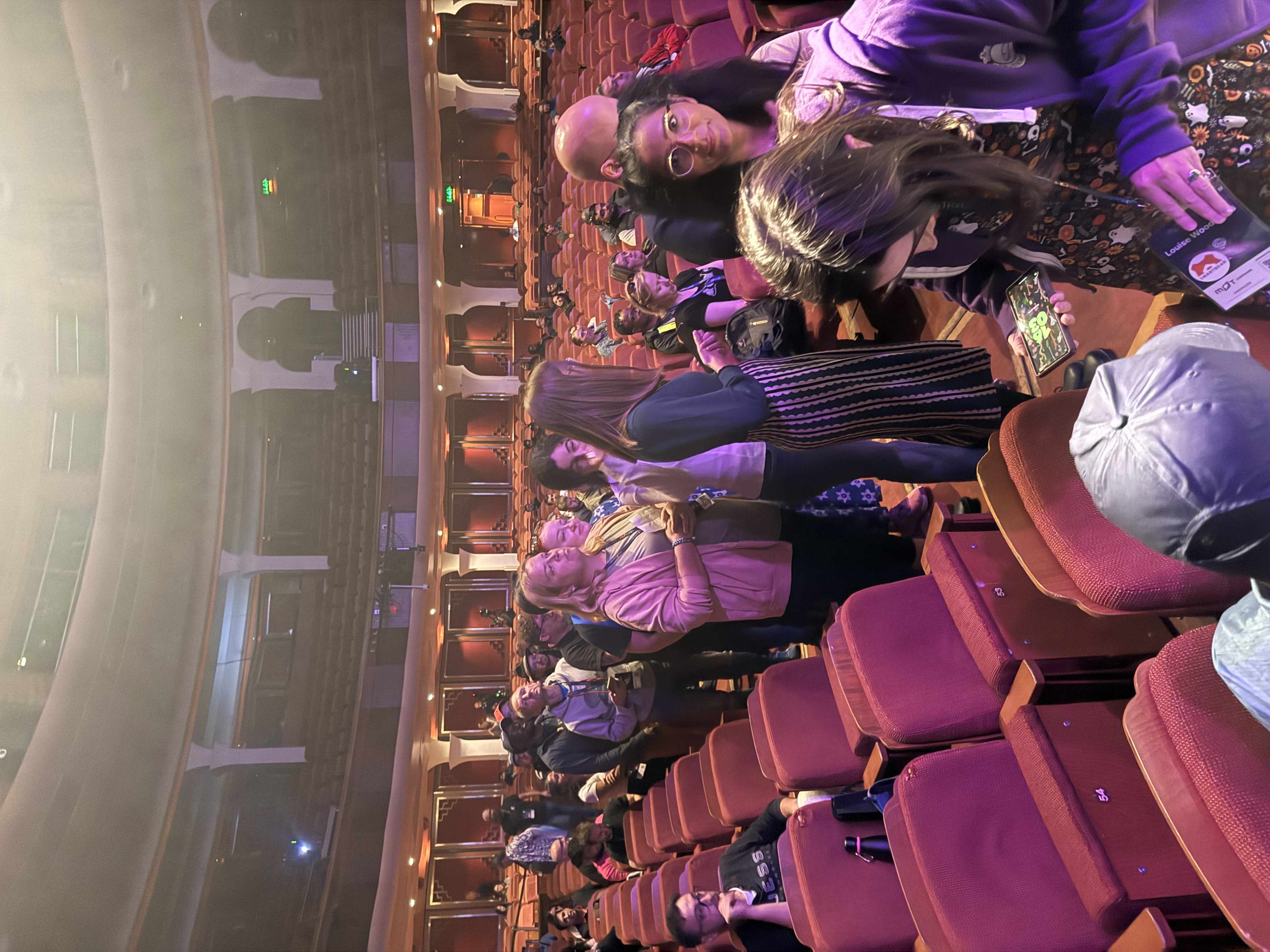 Audience members stand and mill about in the aisles of a large, ornate theatre with red velvet seats before joining the stage (out of shot). 
