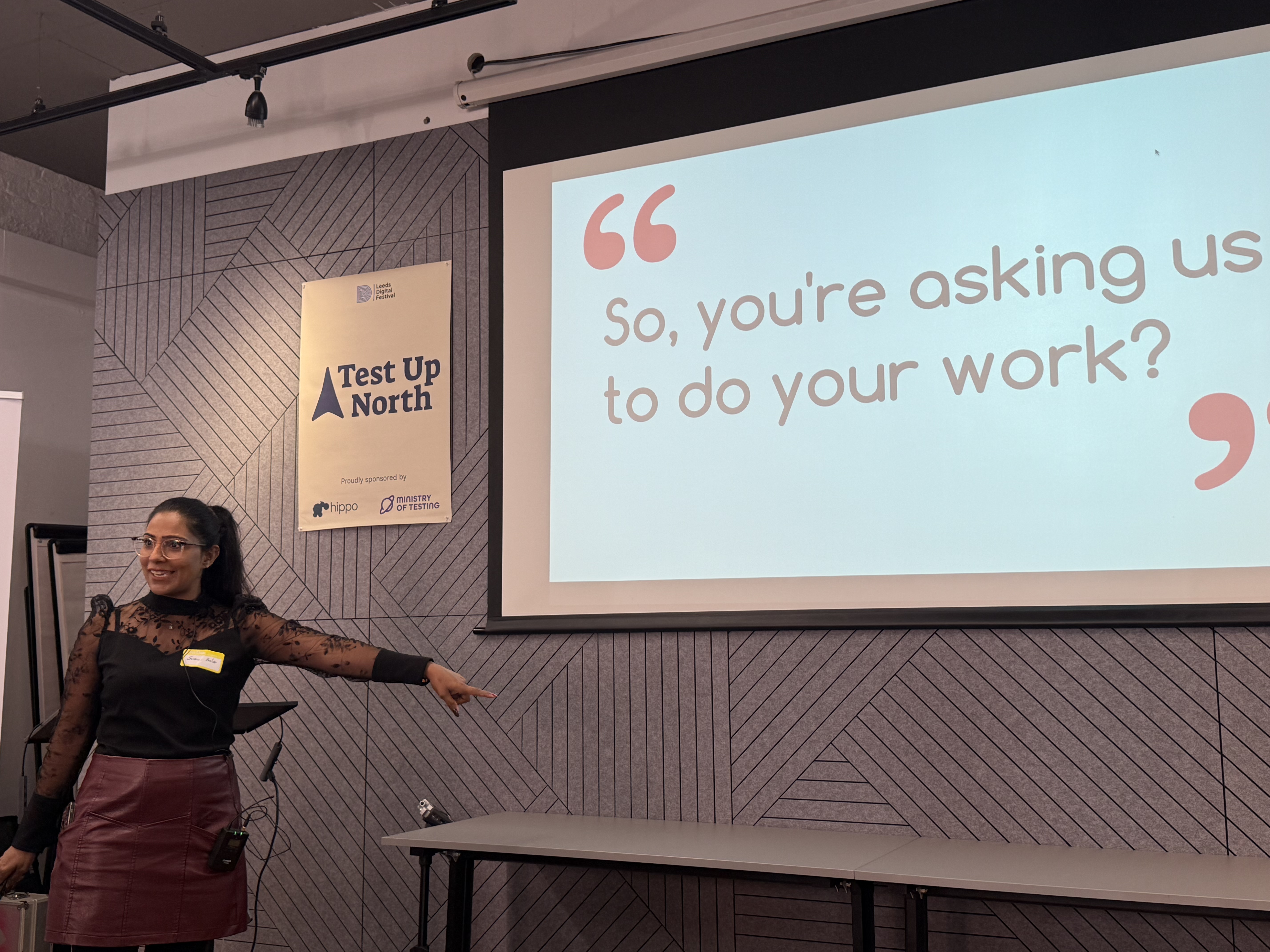 Suman presenting wearing a black top and red leather skirt. The screen shows the quote, ‘So, you’re asking us to do your work?’