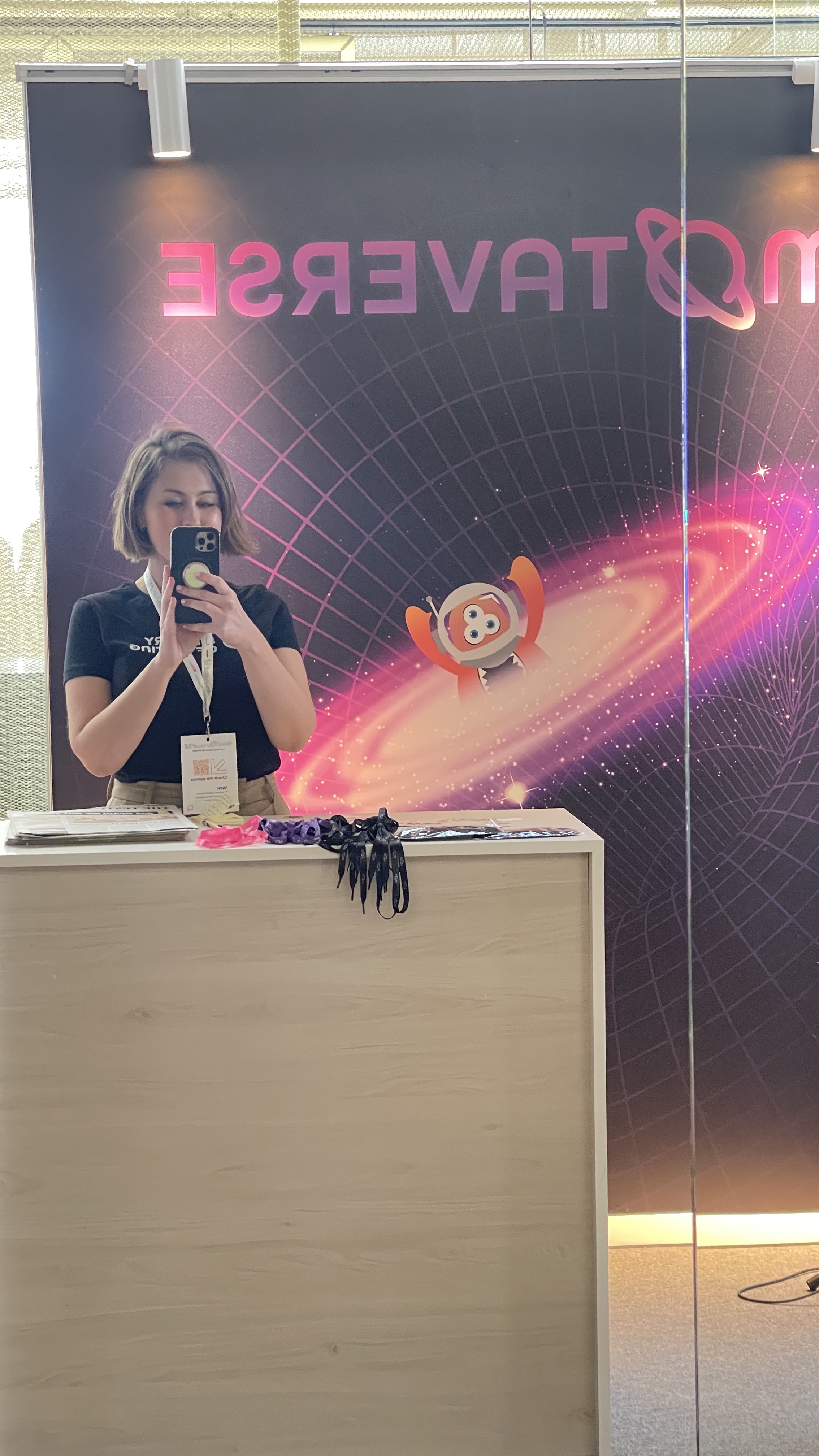 mirror selfie at the Ministry of Testing stand before opening on Day 1 of Selenium Conf 2025. The MoTaverse banner with the mascot and purple galaxy design is visible behind her, with lanyards and materials laid out on the desk.