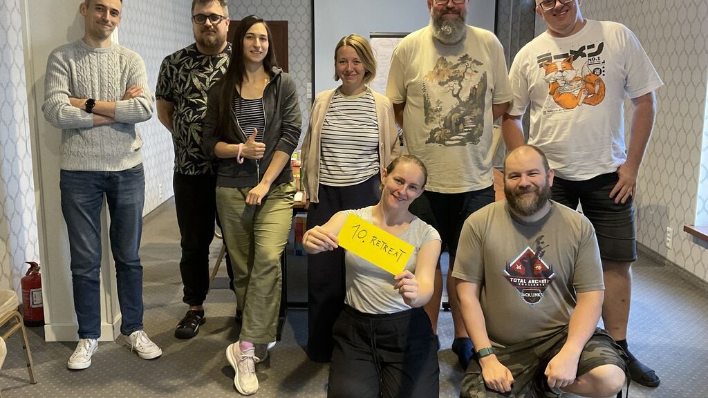 A group of eight people in a conference room. Six standing, two crouching. A whiteboard with notes in the background. One of the two crouching persons is holding a sign with the text "10 RETREAT"