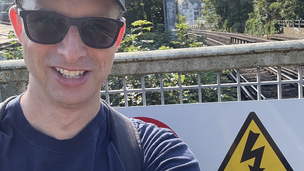 A man wearing a black cap and blue t-shirt with a big character on it stands at the end of a railway platform. 
