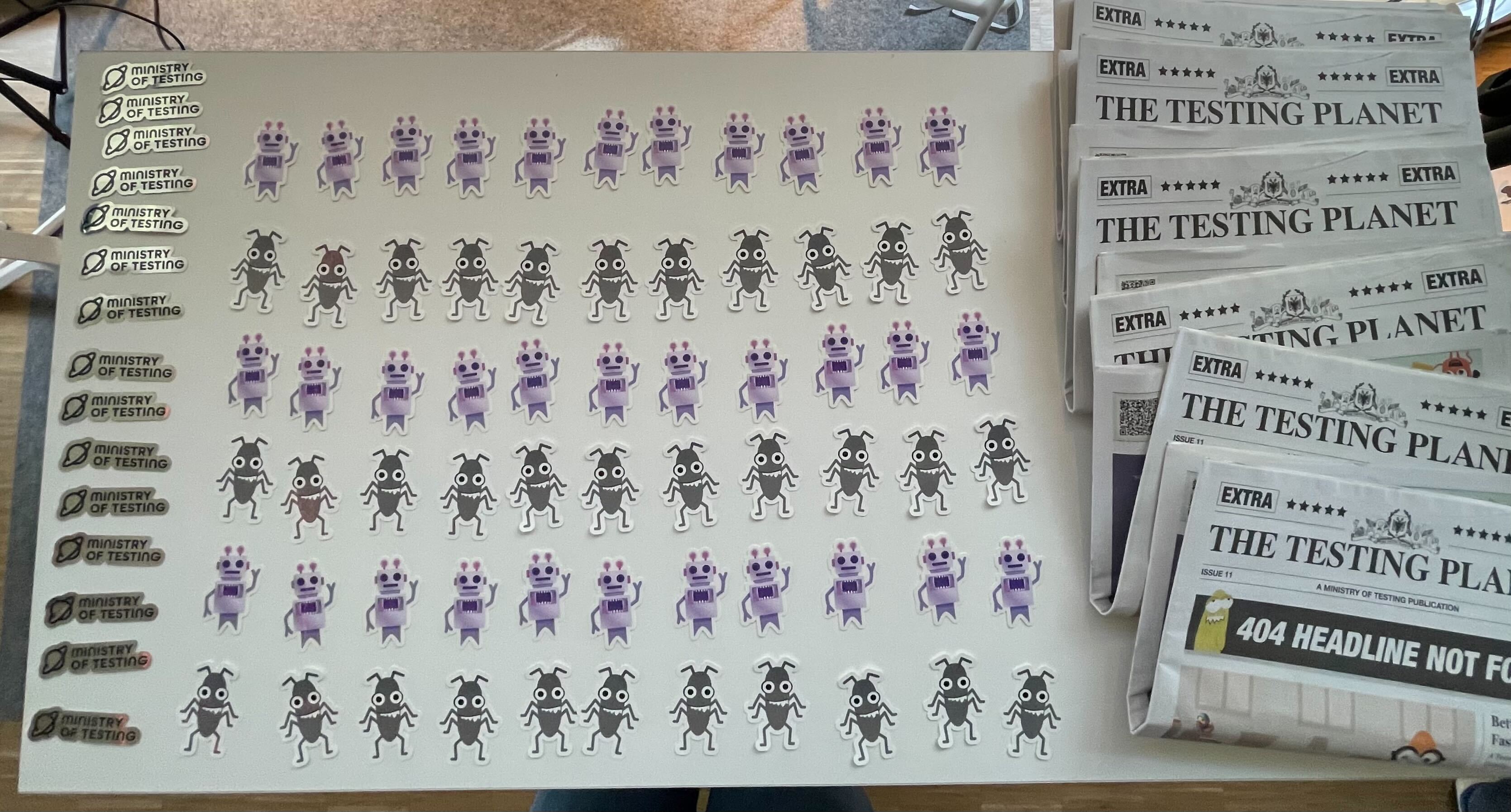 Ministry of Testing, Bug and Automator stickers laid out on a table alongside printed copies of The Testing Planet.