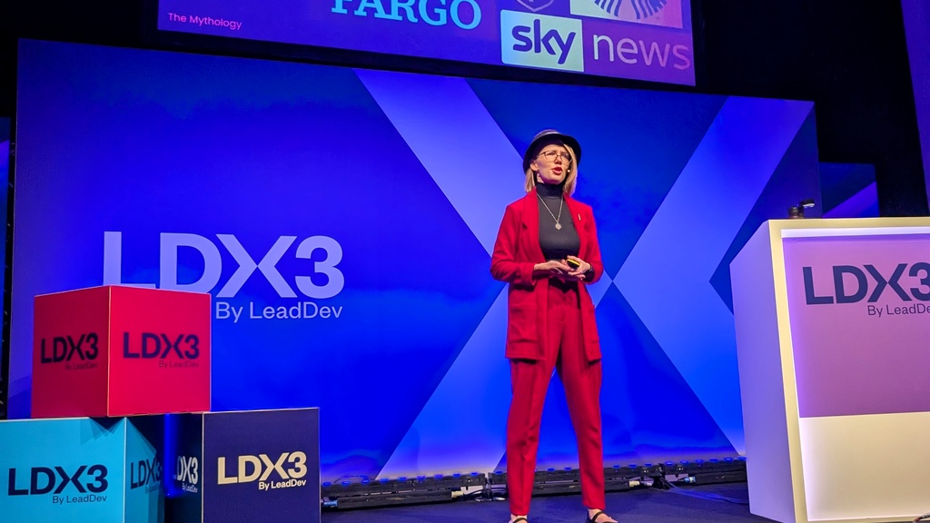 Christine standing on the LDX3 Stage. She is wearing a red suit and blue hat. Behind her is her slide showing the number '5.6 billion' and showing a bunch of logos of fortune 500 companies which lost in total 5.6 billion dollars through the global outage caused by Crowdstrike