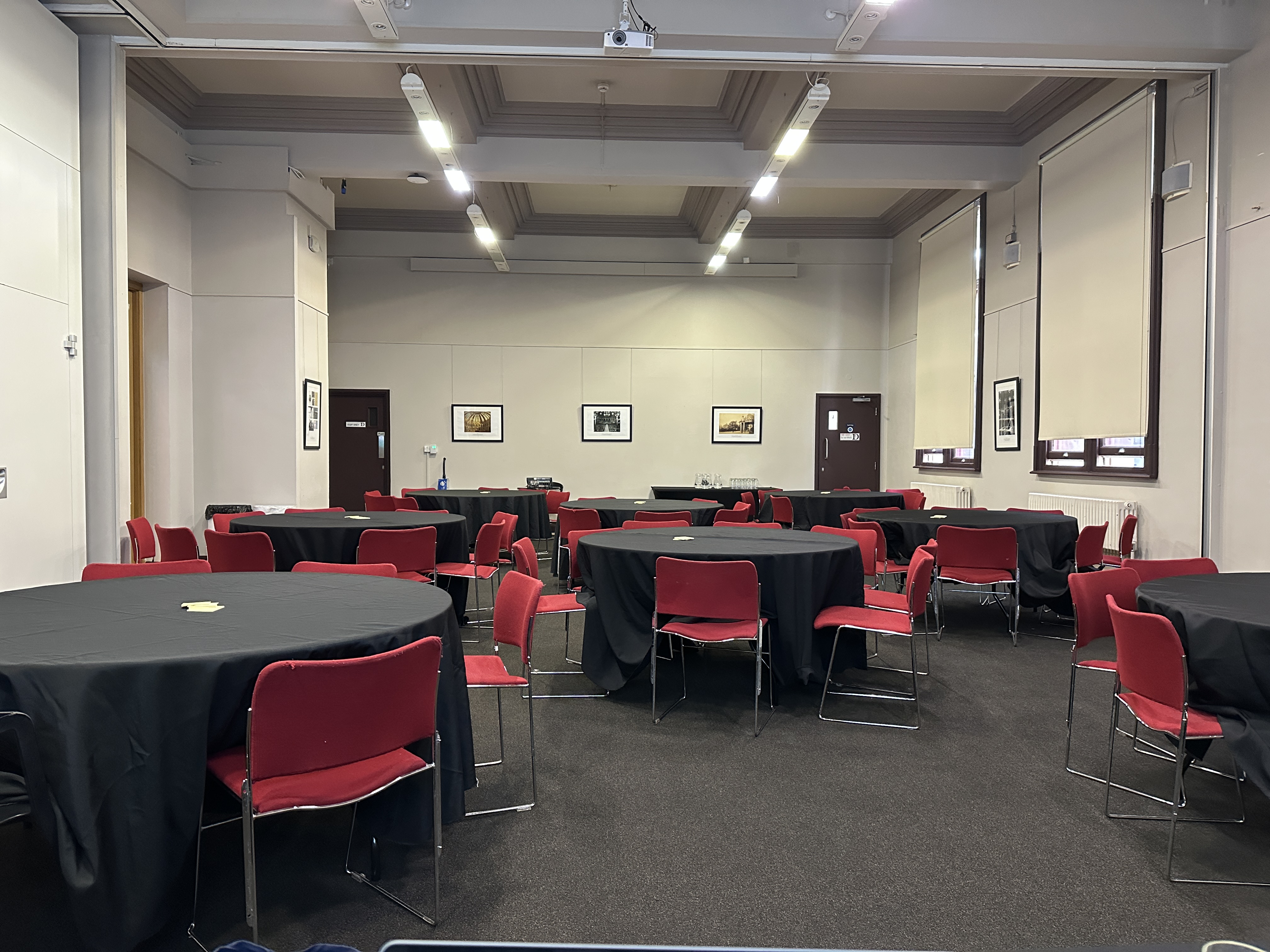 The Founder’s Room at the Brighton Dome, set up for an event. The room features round tables covered with black tablecloths and surrounded by red chairs. The walls are light-coloured with framed photographs, tall windows with blinds, and overhead strip lighting creating a bright, professional atmosphere.
