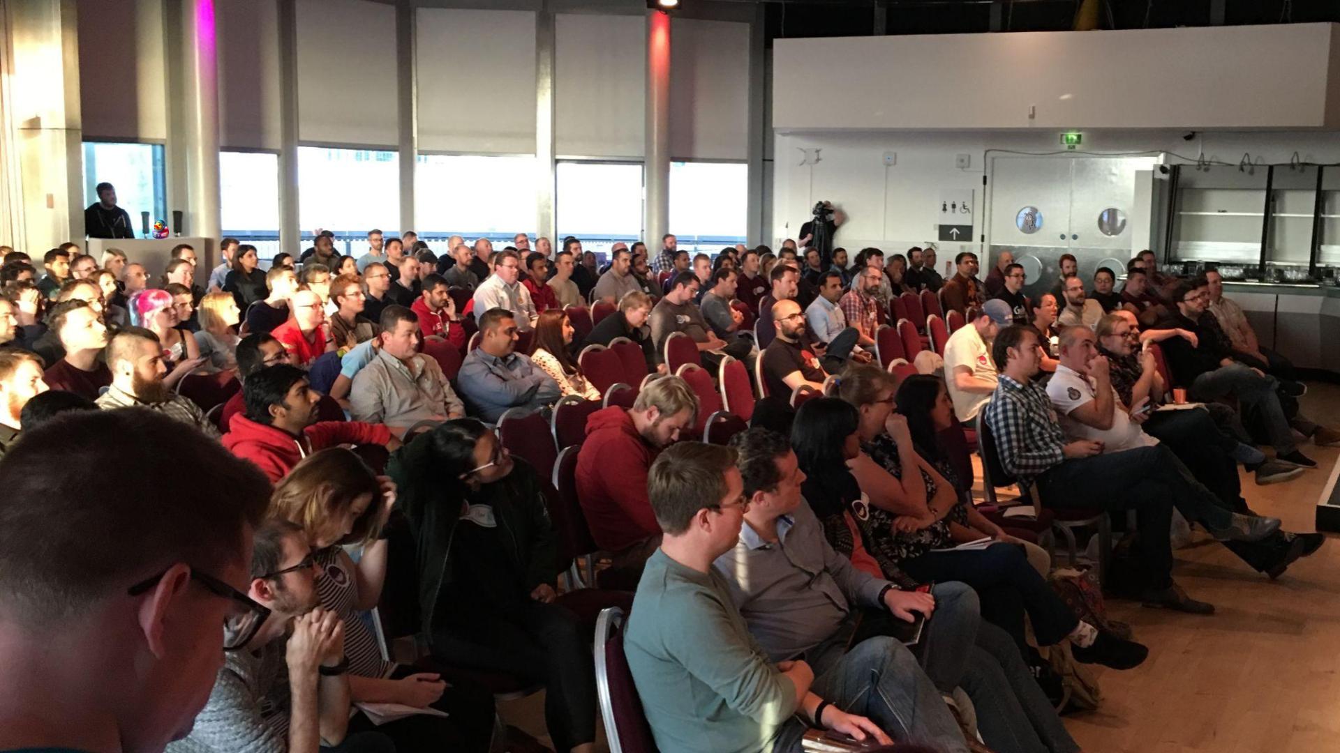 A large group of people face the stage in a conference room space. They appear interested. A duck and bug character have been added to the photo and appear to be hiding.