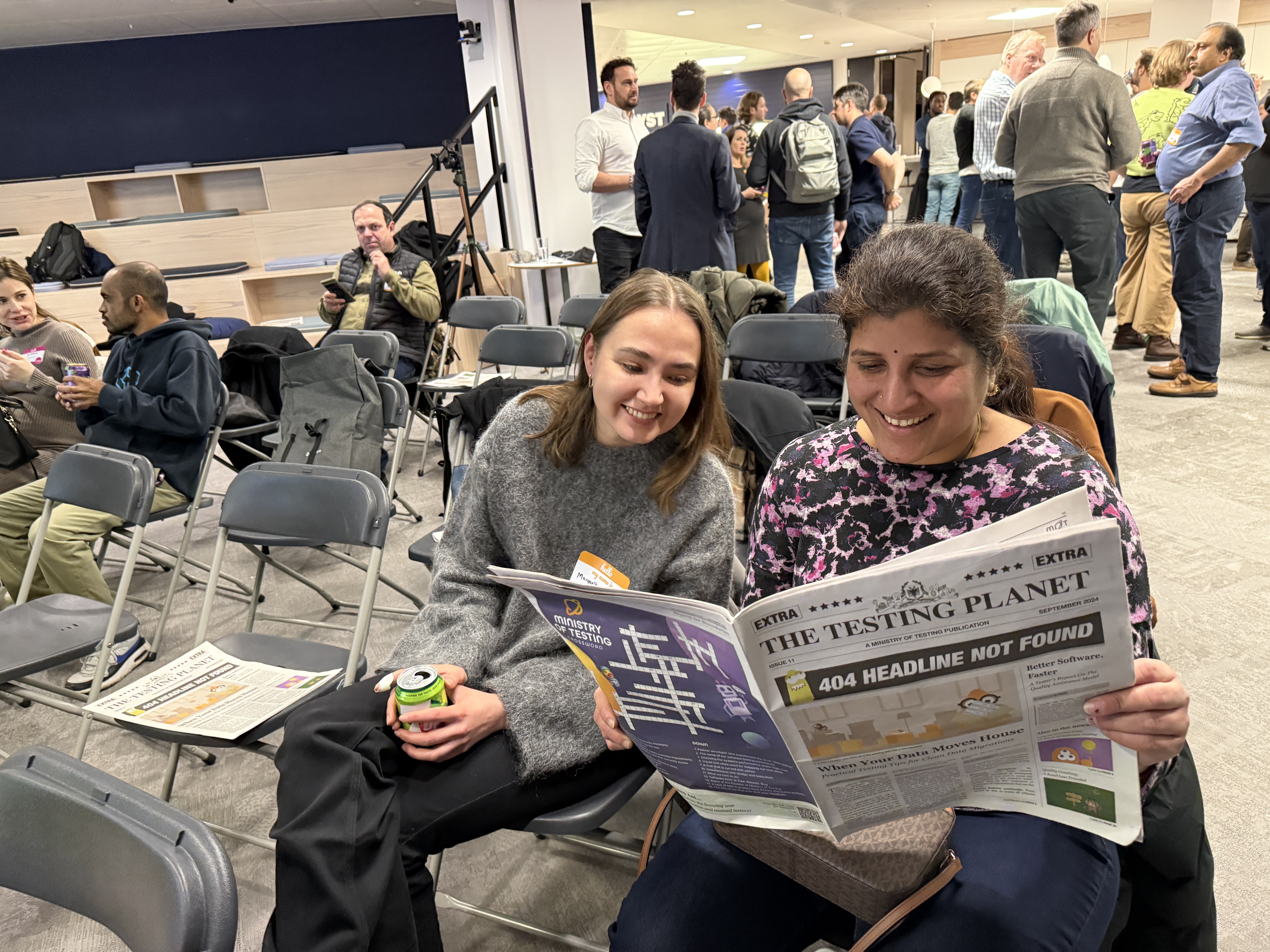 Two women sat on a row of chairs reading a newspaper. People are standing in the background in an open office space. Lots of chat is happening.  