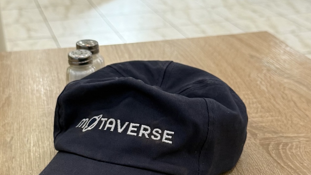 A blue baseball cap sits on a table with MoTaverse on it. In the background are serving tables with breakfast items on.  image
