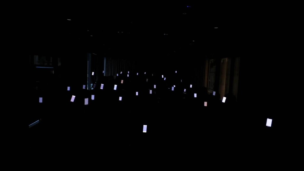 A conference audience raising illuminated smartphones, each displaying coordinated colours and patterns as part of a ... image