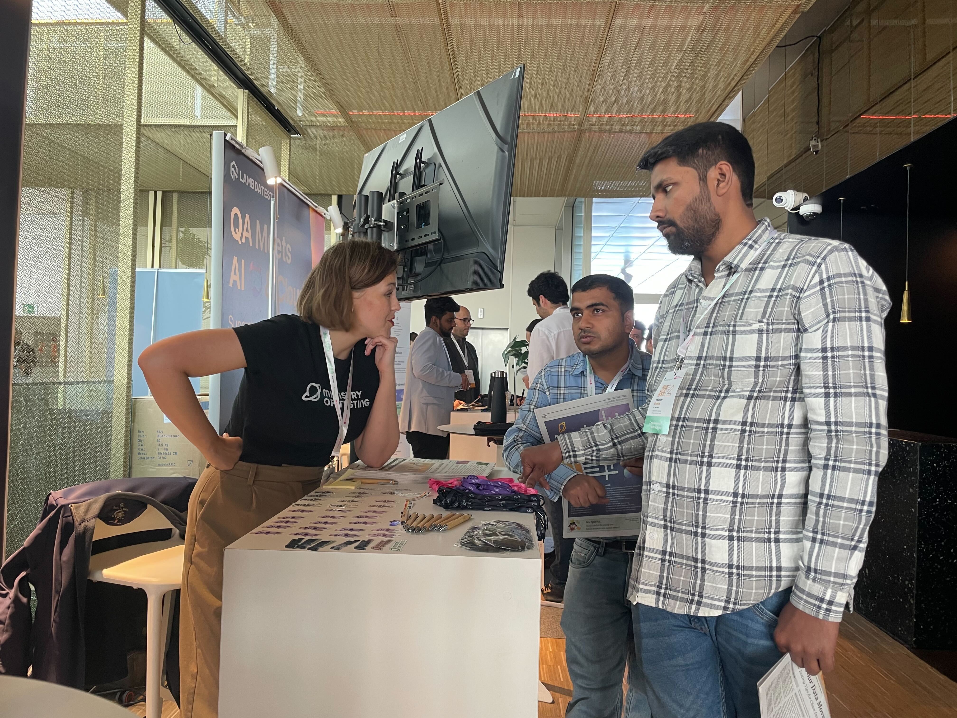 Alexandra leans in at the Ministry of Testing booth and talks to two people about the MoTaverse.