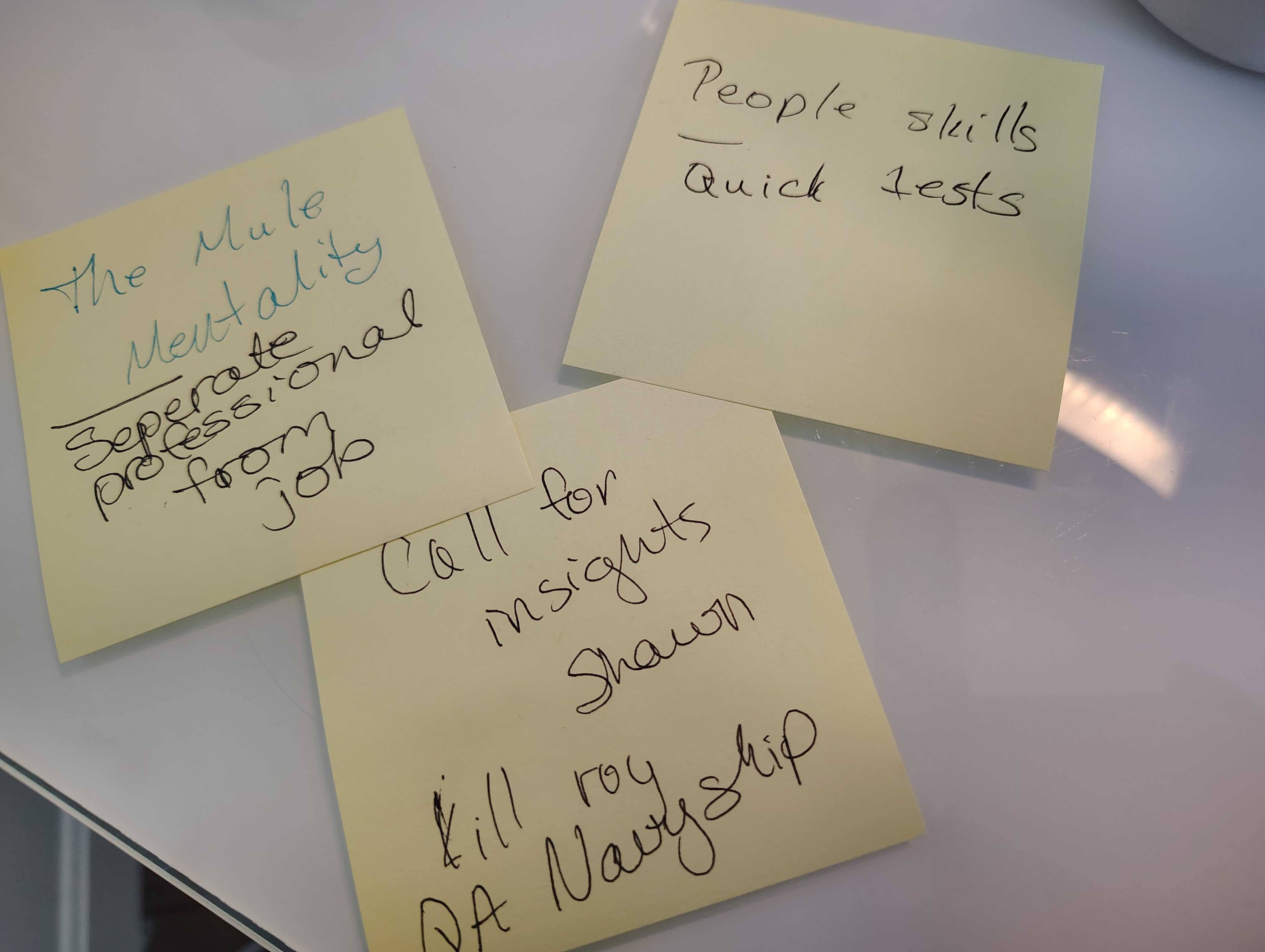 Alt text: Three yellow sticky notes on a white desk with handwritten notes. One reads “The Mule Mentality” with “separate professional from job”. Another says “People skills” and “Quick tests.” The third reads “Call for insights Shawn” and “Kilroy QA Navy ship.”
