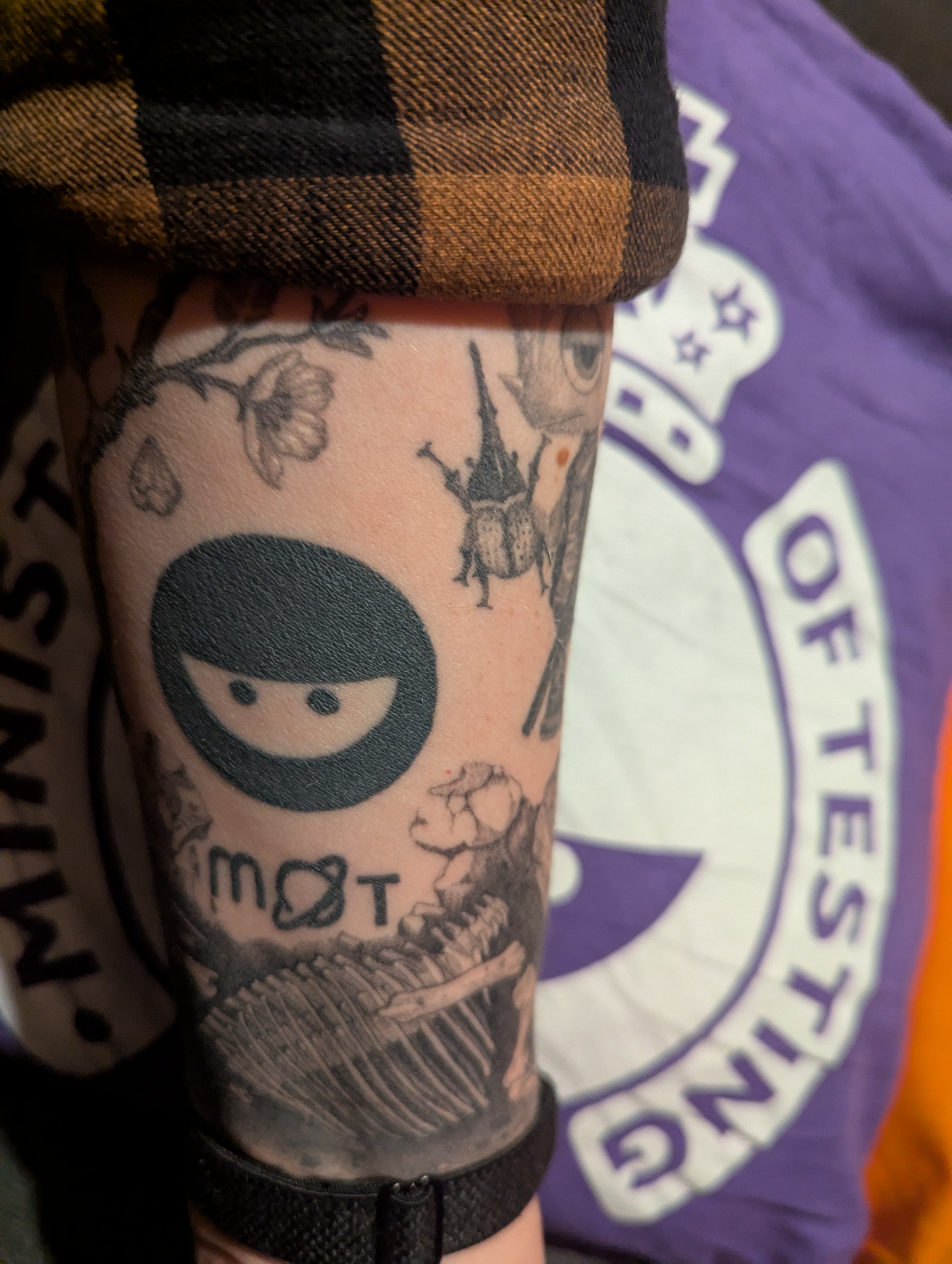 Photo of an arm with both the ministry of testing ninja and the newer mot logo amongst other tattoos.