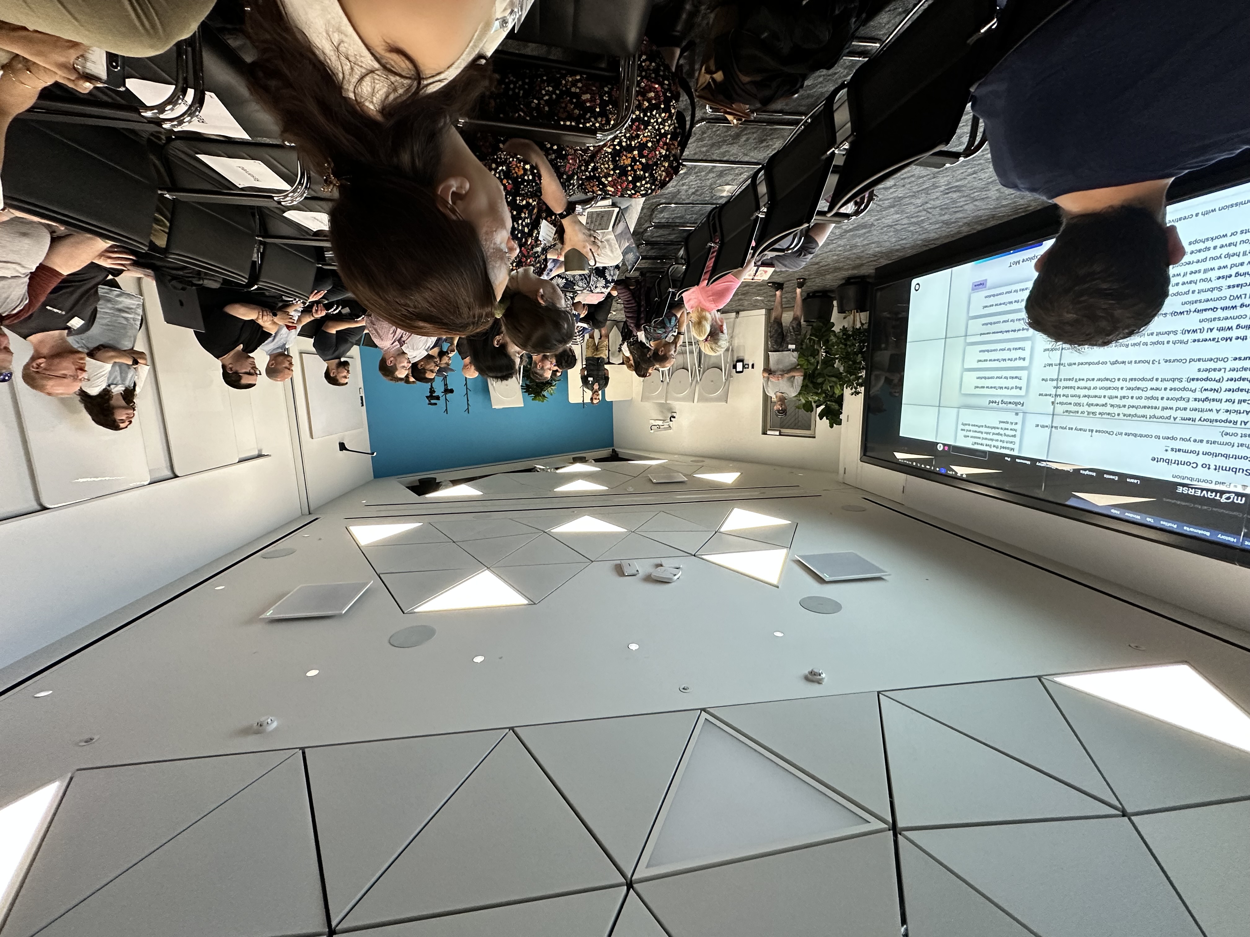 A group of people seated in a modern meeting room watch a presentation on a large screen, while a speaker stands at t... image