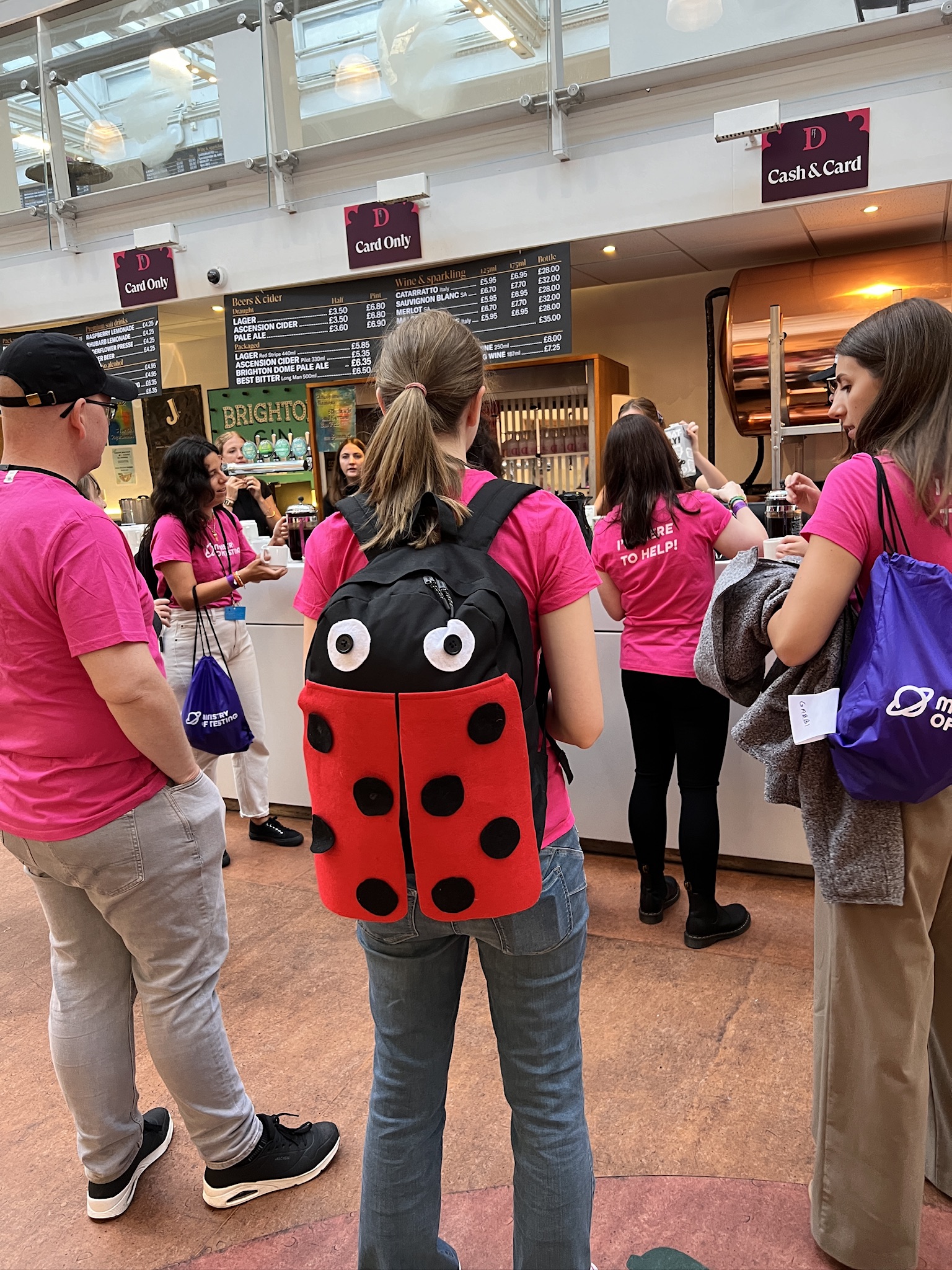 Emily, who is in the middle of the image facing away from the camera has her light hair tied up and is wearing a DIY bug bag. She stands next to Rich and Gabbi in the Brighton Dome before event guests arrive in September 2024.