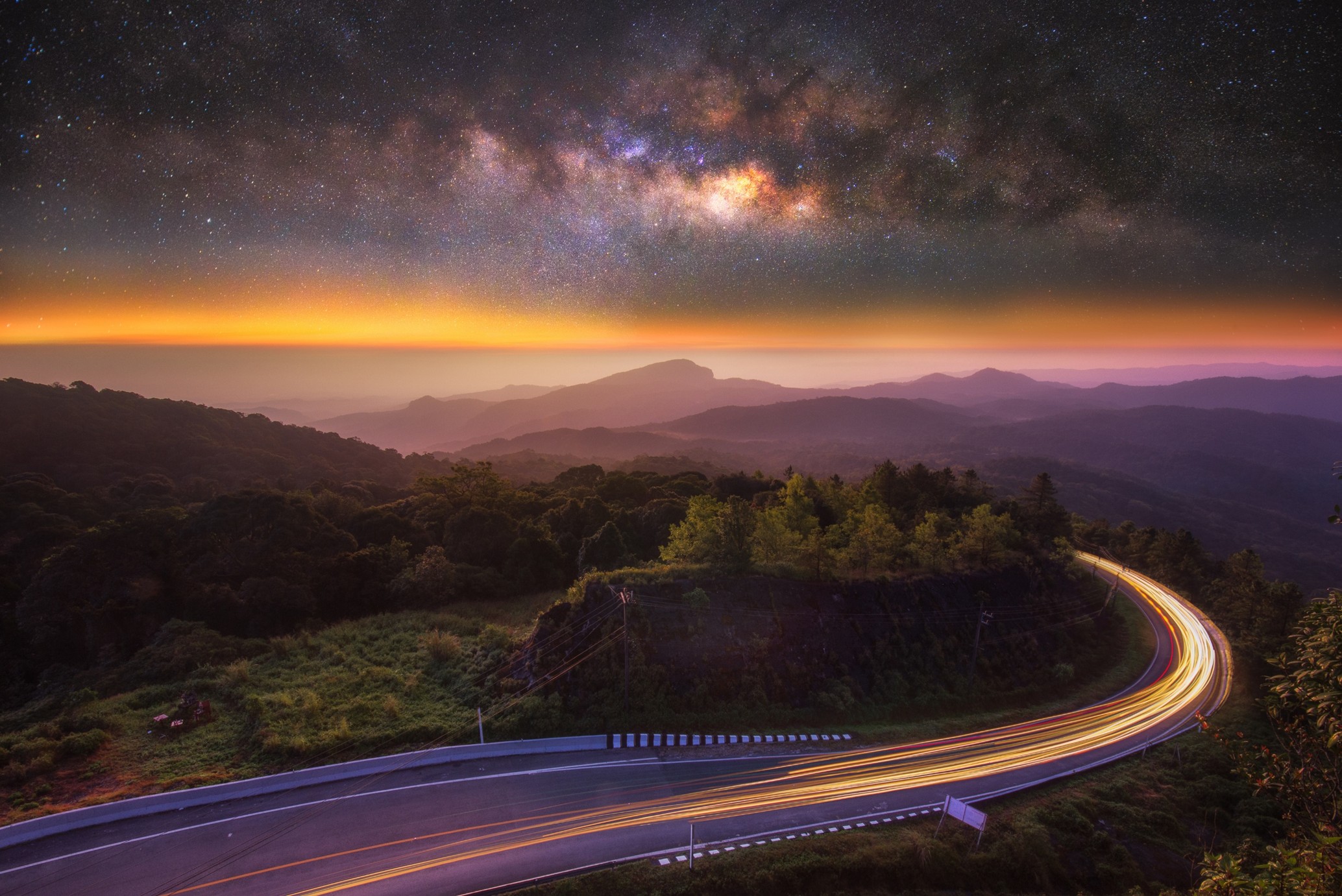 A landscape image with a long exposure of vehicle lights on a bend in the road heading towards mountains and the starry night sky.