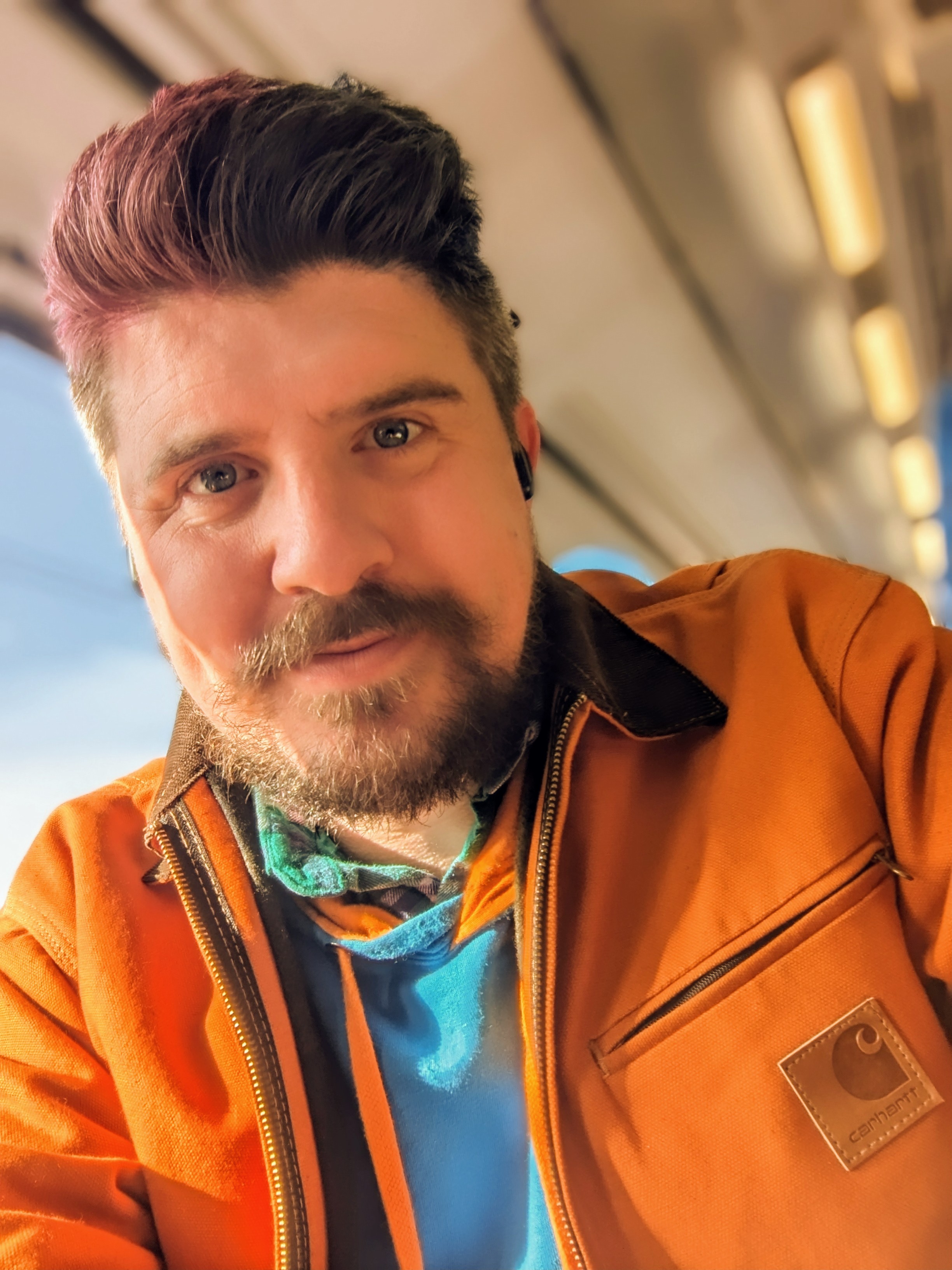 Head and shoulder picture of Scott on a train , wearing a brown jacket, With sunset filter and pink and blue hair fresh for test bash 