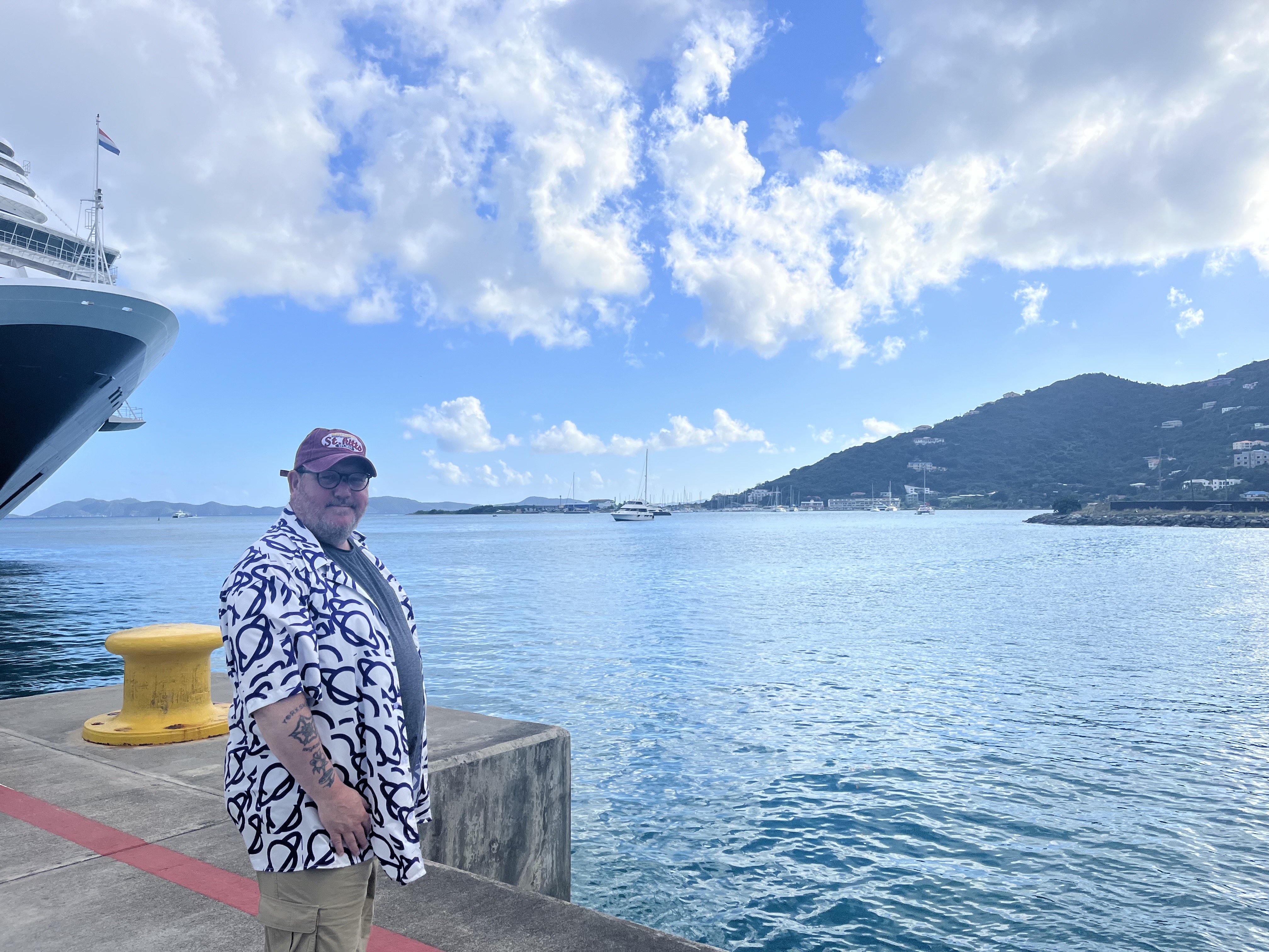 Ady Stokes wearing an MoT planet logo on the dock at the British Virgin Isles. There’s the from of a cruise ship in the background with the bay stretching across to a hillside. 