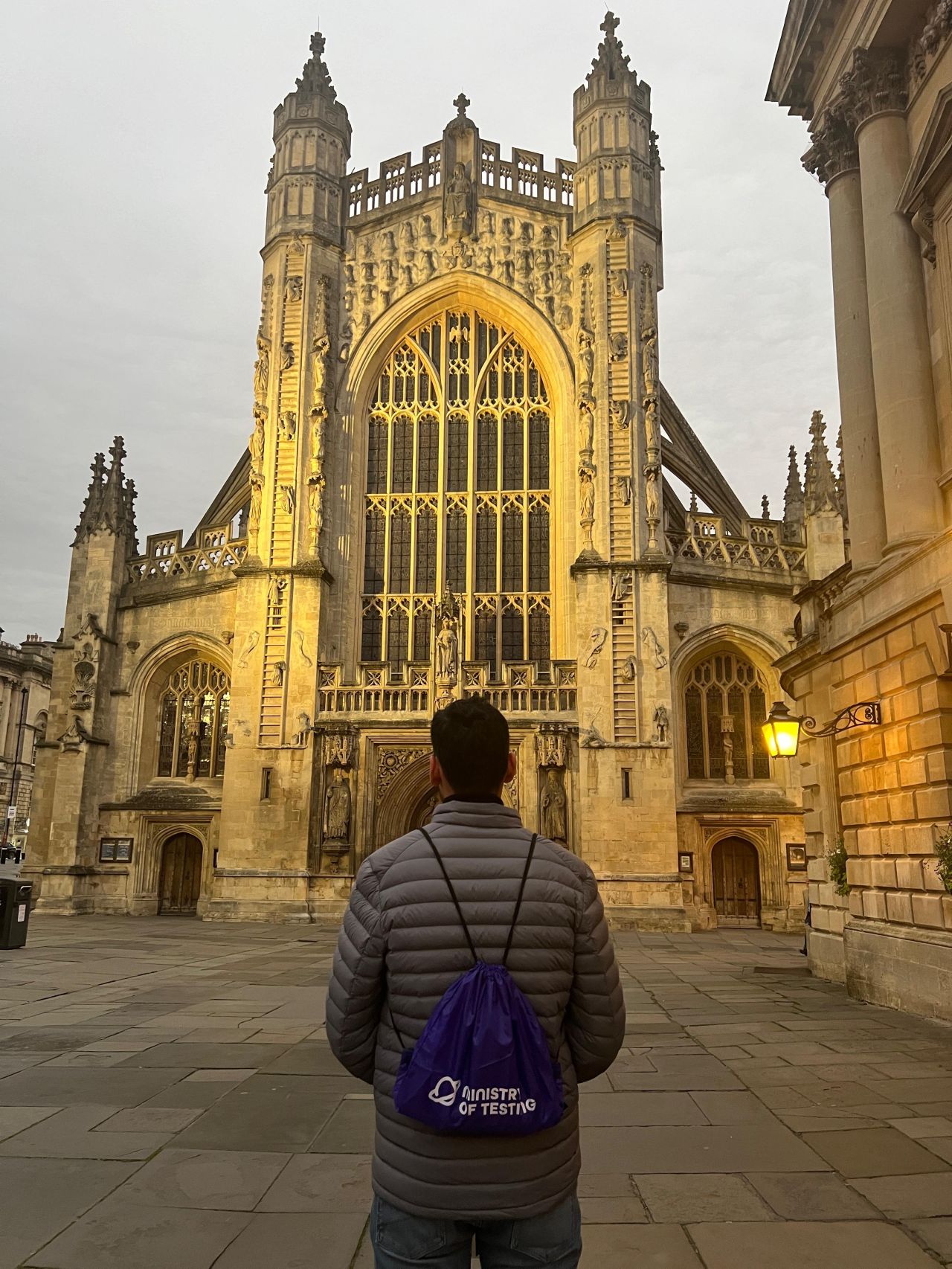 A Man wearing Ministry of testing bag pack, facing the Bath Abbey as the Sun sets over it.