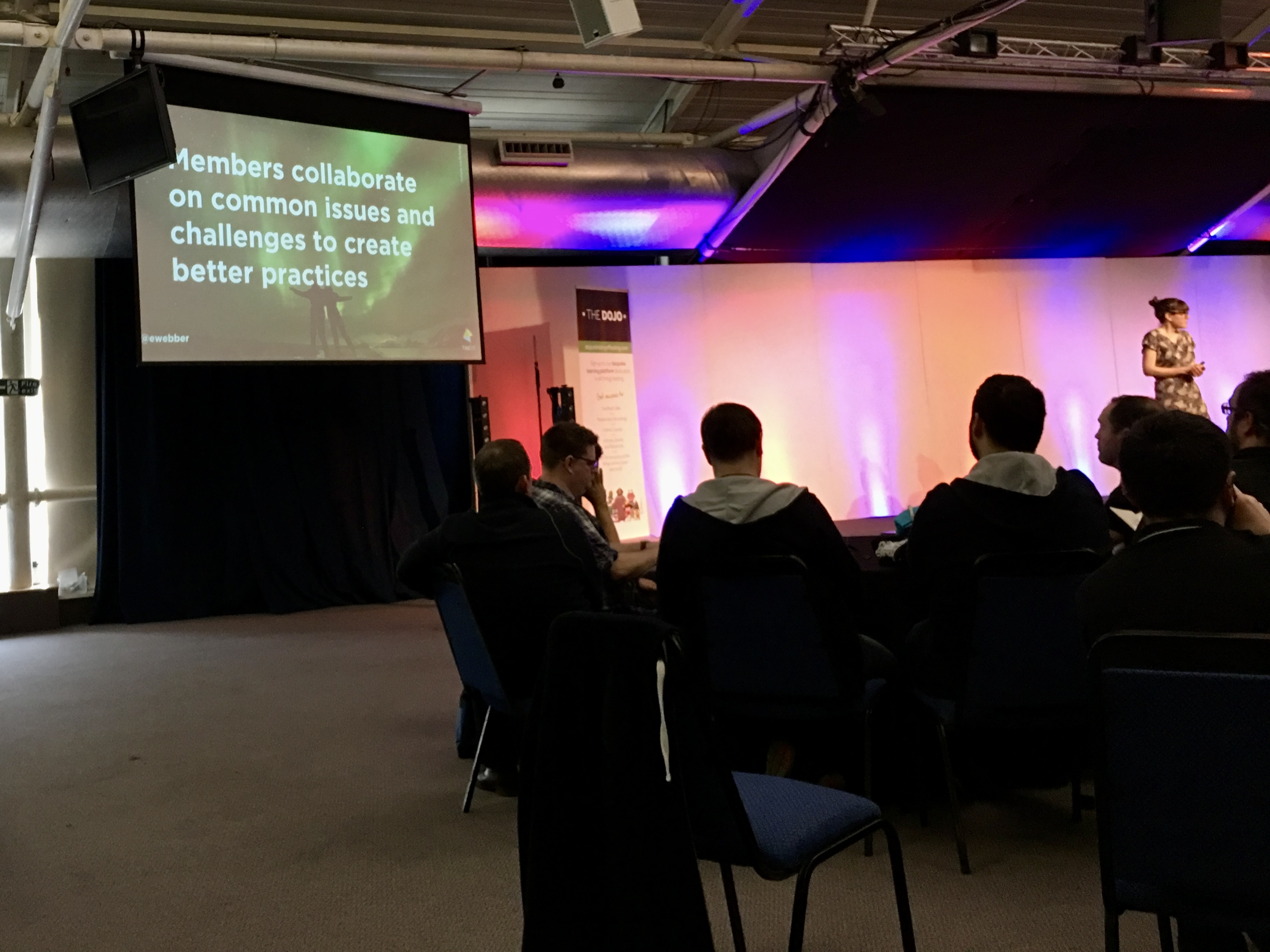 A woman on stage in a large event space. The slide on the screen reads: Members collaborate on common issues and chal... image