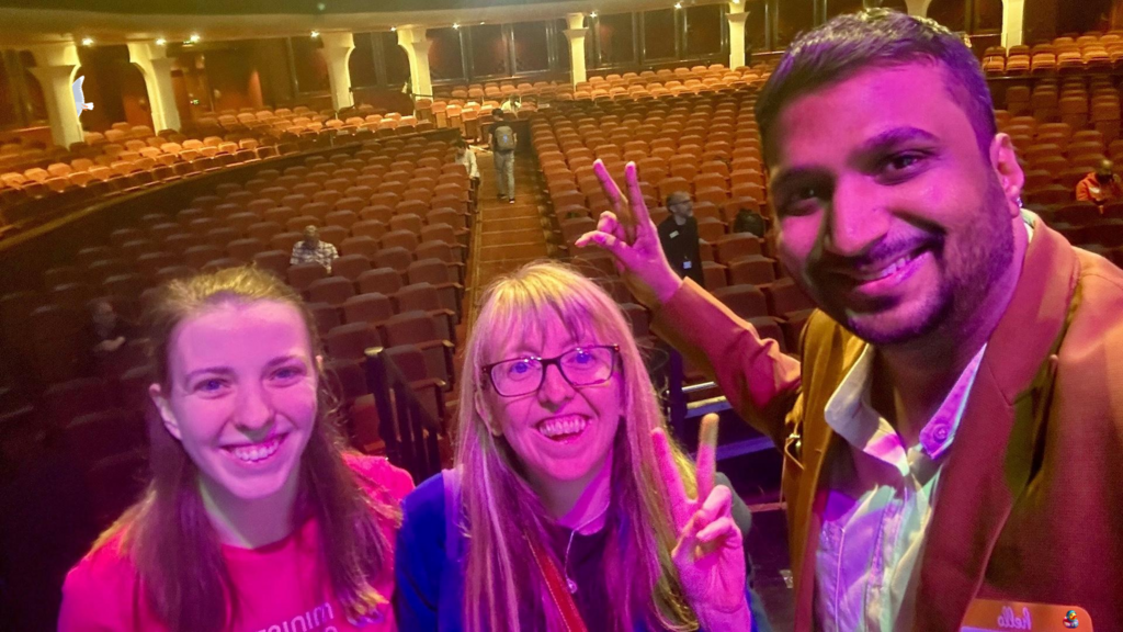 
A man and two women take a selfie in an auditorium with rows of empty seats behind them. The man on the right has dark hair and a beard, is smiling, and is wearing a brown jacket over a light-colored collared shirt. He is holding up two fingers in a peace sign with his left hand. The woman in the middle has blonde hair and glasses, is smiling, and is also holding up two fingers in a peace sign. The woman on the left has dark hair and is smiling. The auditorium seats are red and appear to be empty, except for a few people visible in the distance. A bug, a seagul and a duck character have been added to the image. 