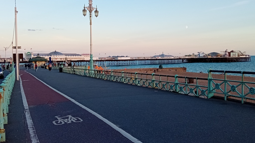 A photograph of the Promenade of Brighton Seafront.  In the picture there is a pavement with a cycle lane.  To the right of the pavement are green railings and a lamp post and then a beach and the sea.  At the back of the photograph is a pier and to the right there is a beach and the sea.  It is dusk and the sky in the picture is clear.
