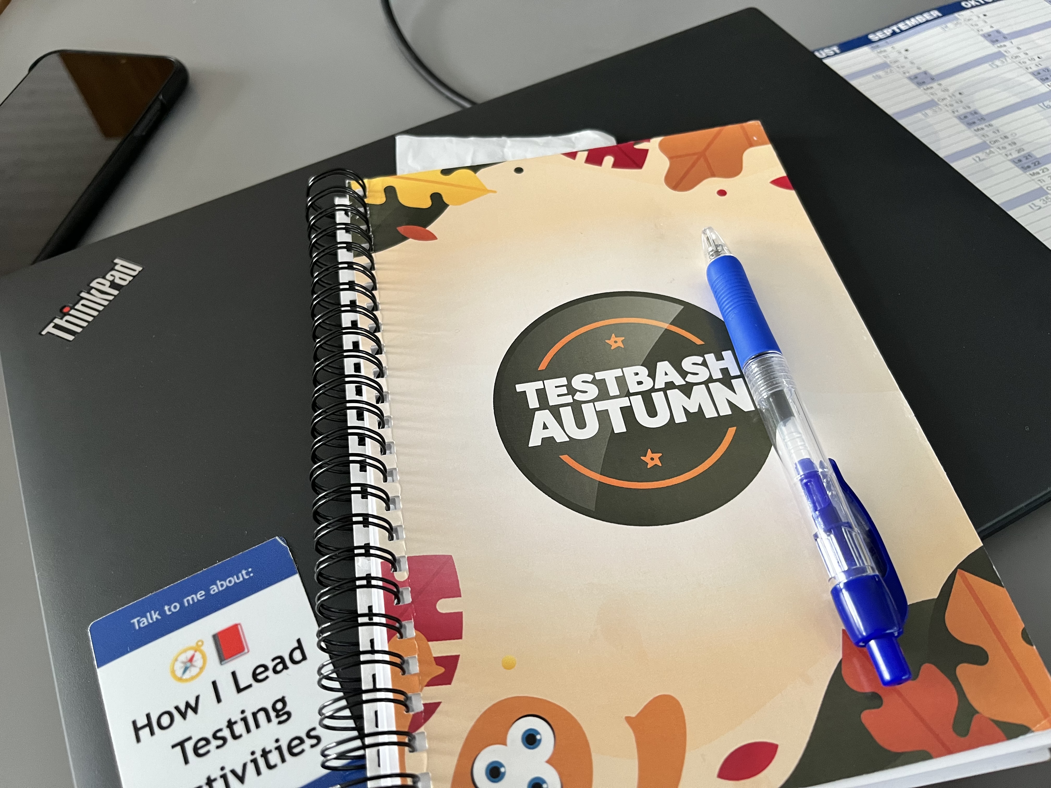 A notebook with the test bash autumn logo placed on a laptop. Also visible a phone, pen and sticker for Leading testing activities 