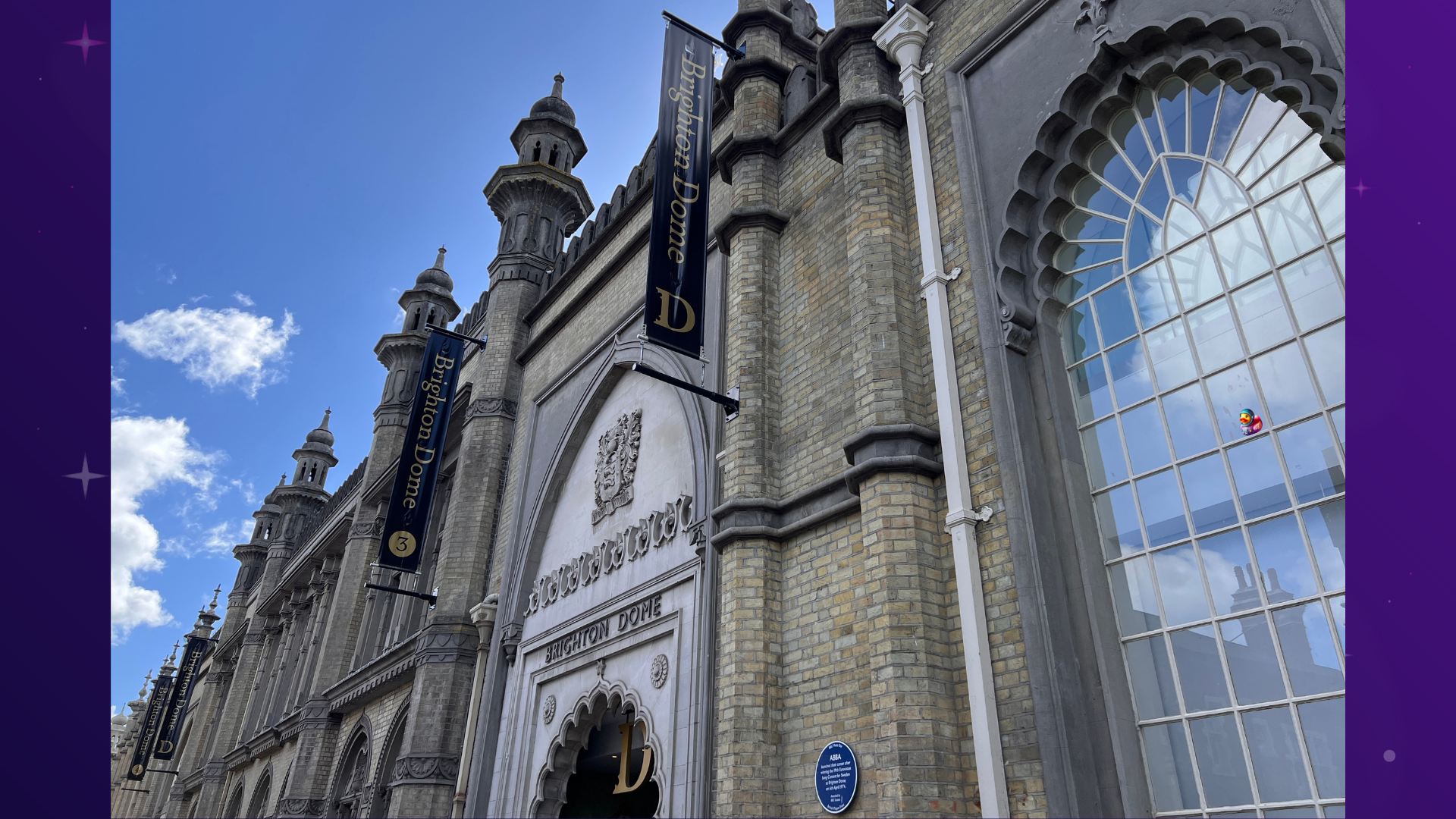The Brighton Dome, a building with a beige brick facade, ornate arches, and intricate stonework, stands against a bright blue sky with scattered clouds. A banner with the "Brighton Dome" logo hangs from the building. A small, colourful, cartoonish bug is visible on the upper-right window. A space duck, a space seagull and a bug character have been added to the image. 