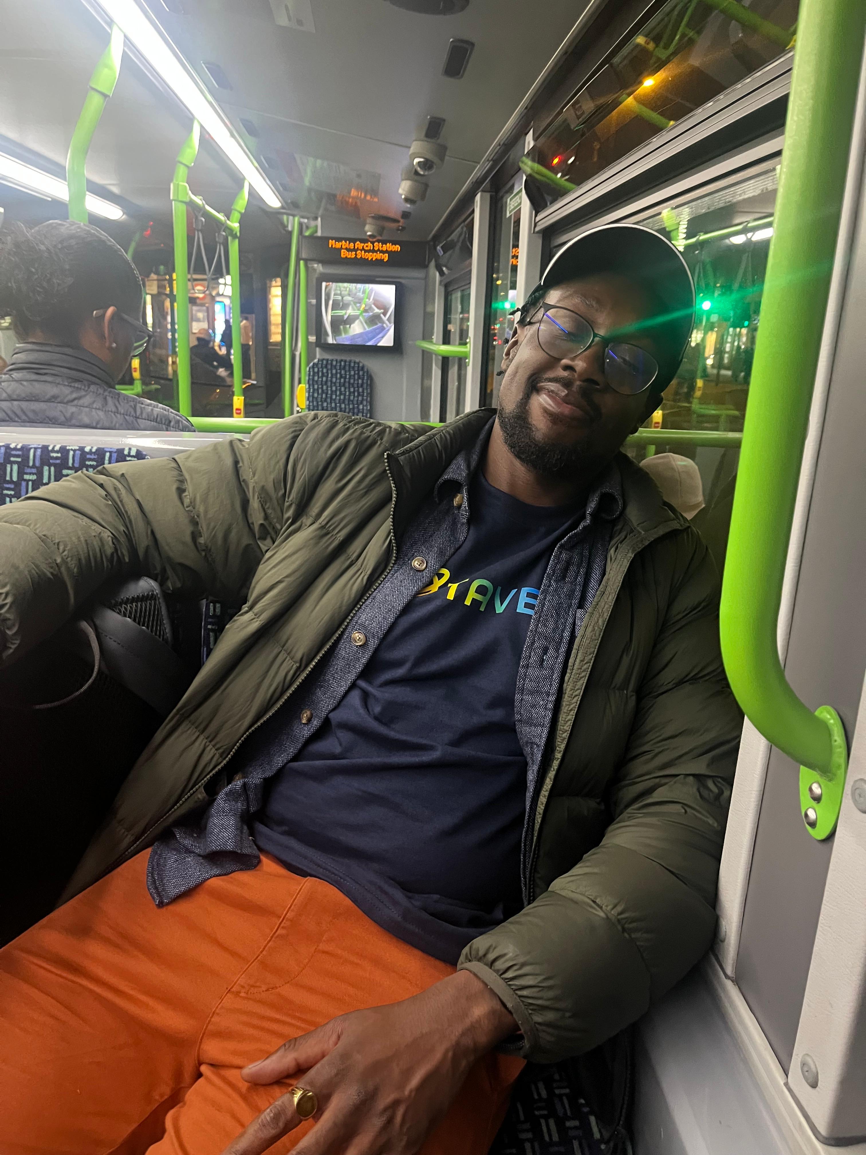 A black man wearing glasses, an olive-green jacket, and orange pants sits on a bus at night, leaning back and looking at the camera. Other passengers are visible in the background.