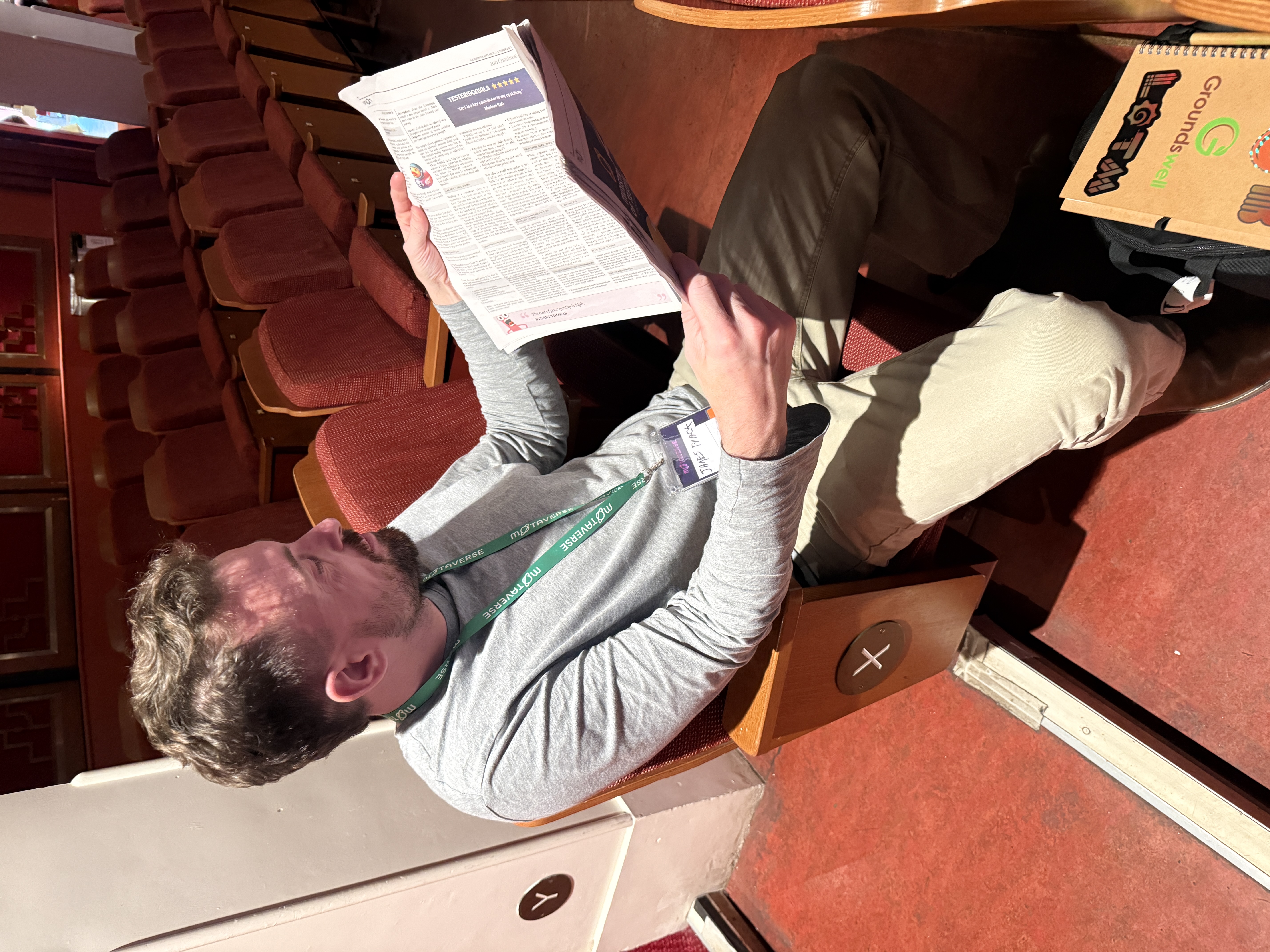 A bearded man with light brown hair sits in a red upholstered theatre seat, reading a newspaper. He is wearing a light gray long-sleeved shirt, khaki trousers, and dark brown boots, with a green lanyard around his neck. The theatre is mostly empty, with rows of similar red seats visible behind him. A notebook or binder rests on the floor next to his feet.