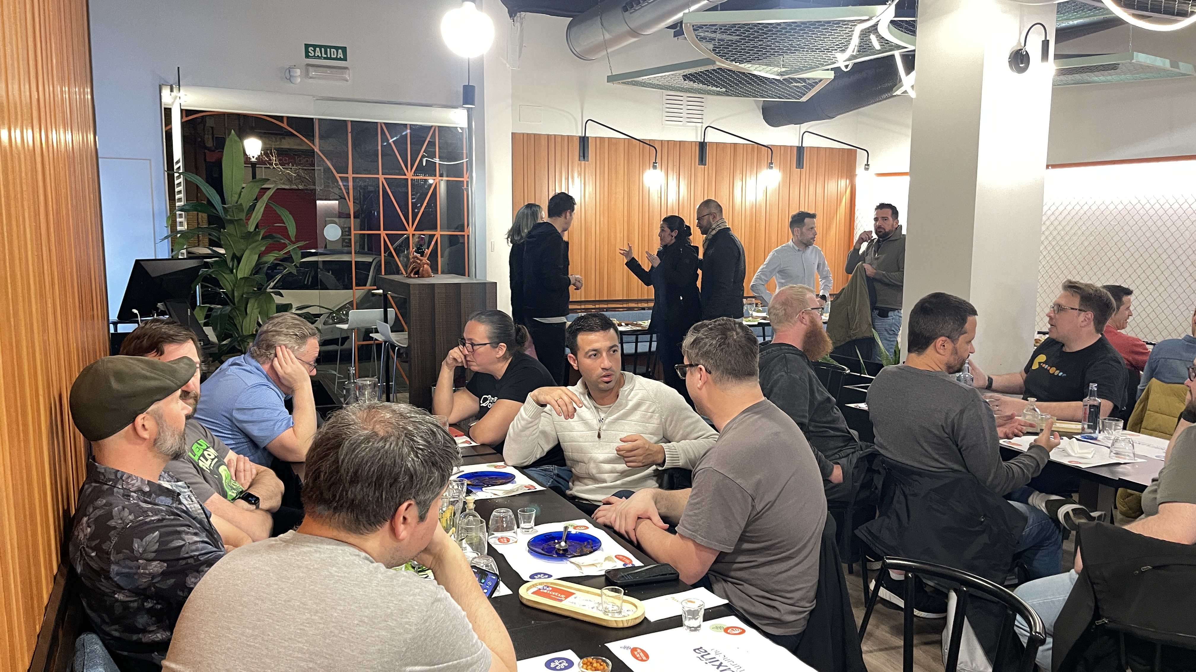 A wide shot of the Selenium Conf 2025 pre-conference speakers' dinner at Maruxiña, with groups deep in conversation. Rosie Sherry and Sergio Olmos are talking at the front table, while other speakers stand and chat near the back of the restaurant.