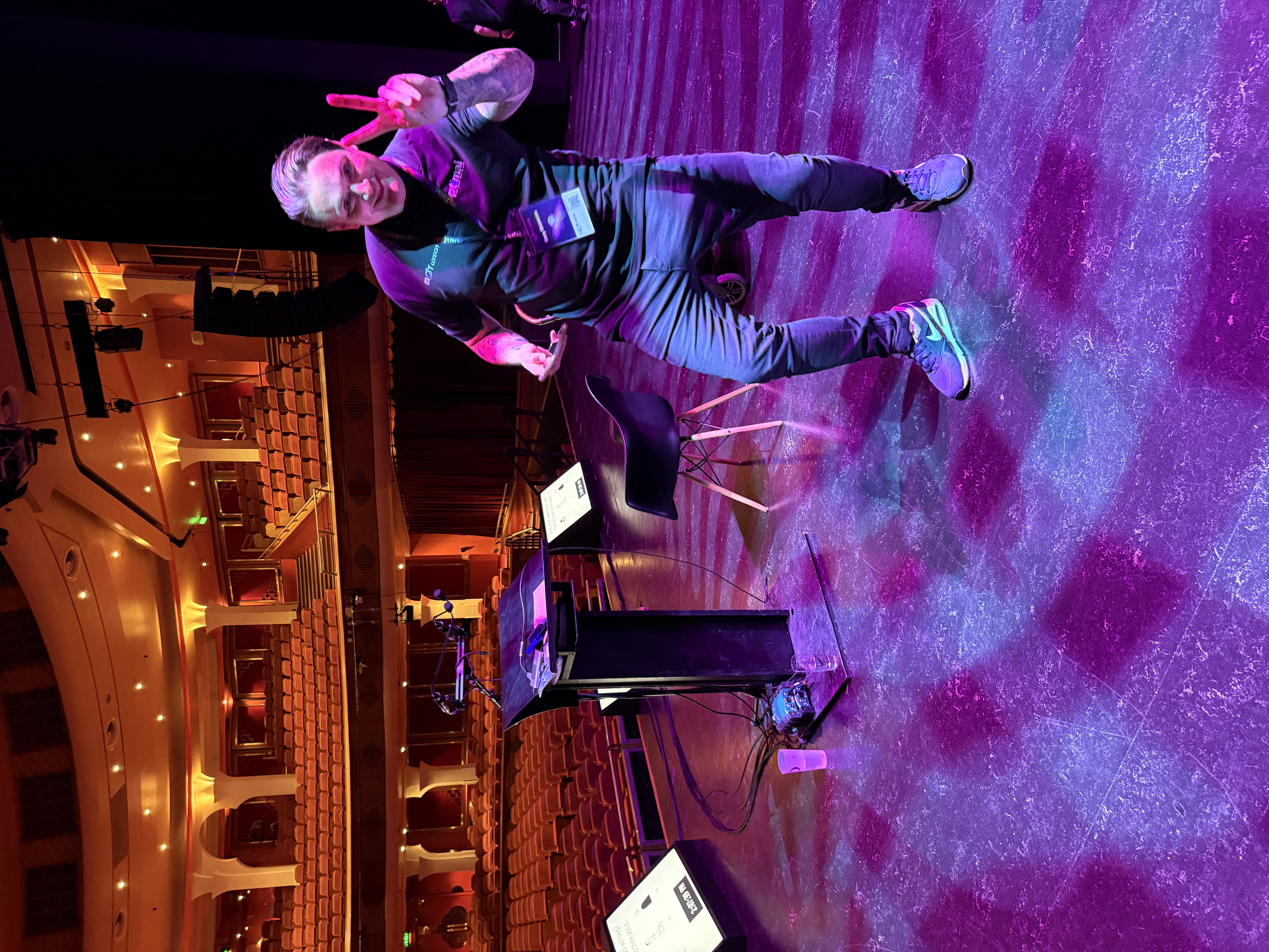 A smiling man stands centre stage in a dimly lit theatre, making a peace sign with his right hand. He is wearing a dark t-shirt, dark jeans, and trainers, with an event badge hanging from a lanyard around his neck. The stage floor is purple and reflects the lighting. Behind him are empty rows of red velvet seats leading up to balconies. A dark lectern with a screen and a black chair are also visible on the stage.
