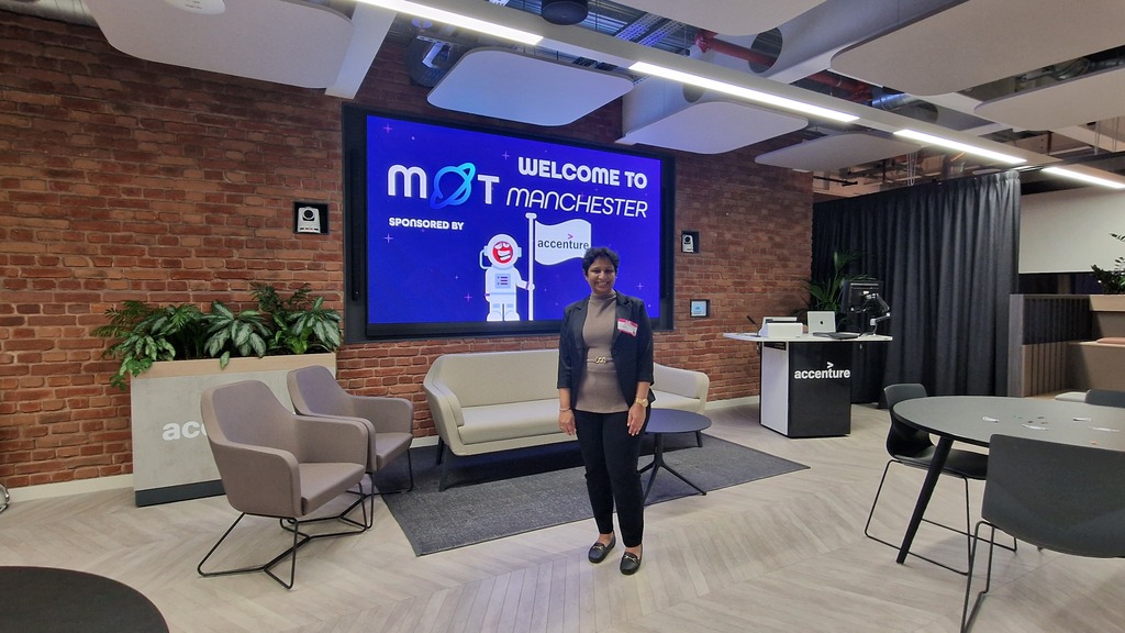 A person stands smiling in the center of a modern, open-plan office lounge. They are wearing a dark blazer over a tan... image